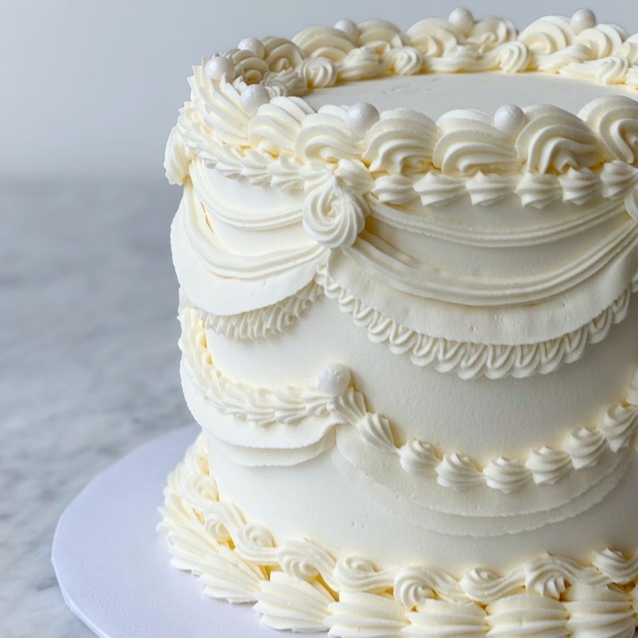A close-up, side-angle view of a tall, monochromatic white cake featuring intricate Lambeth-style piping. Elegant buttercream swags and delicate scalloped borders decorate the sides, accented with small white edible pearls. The cake sits on a white marble surface.