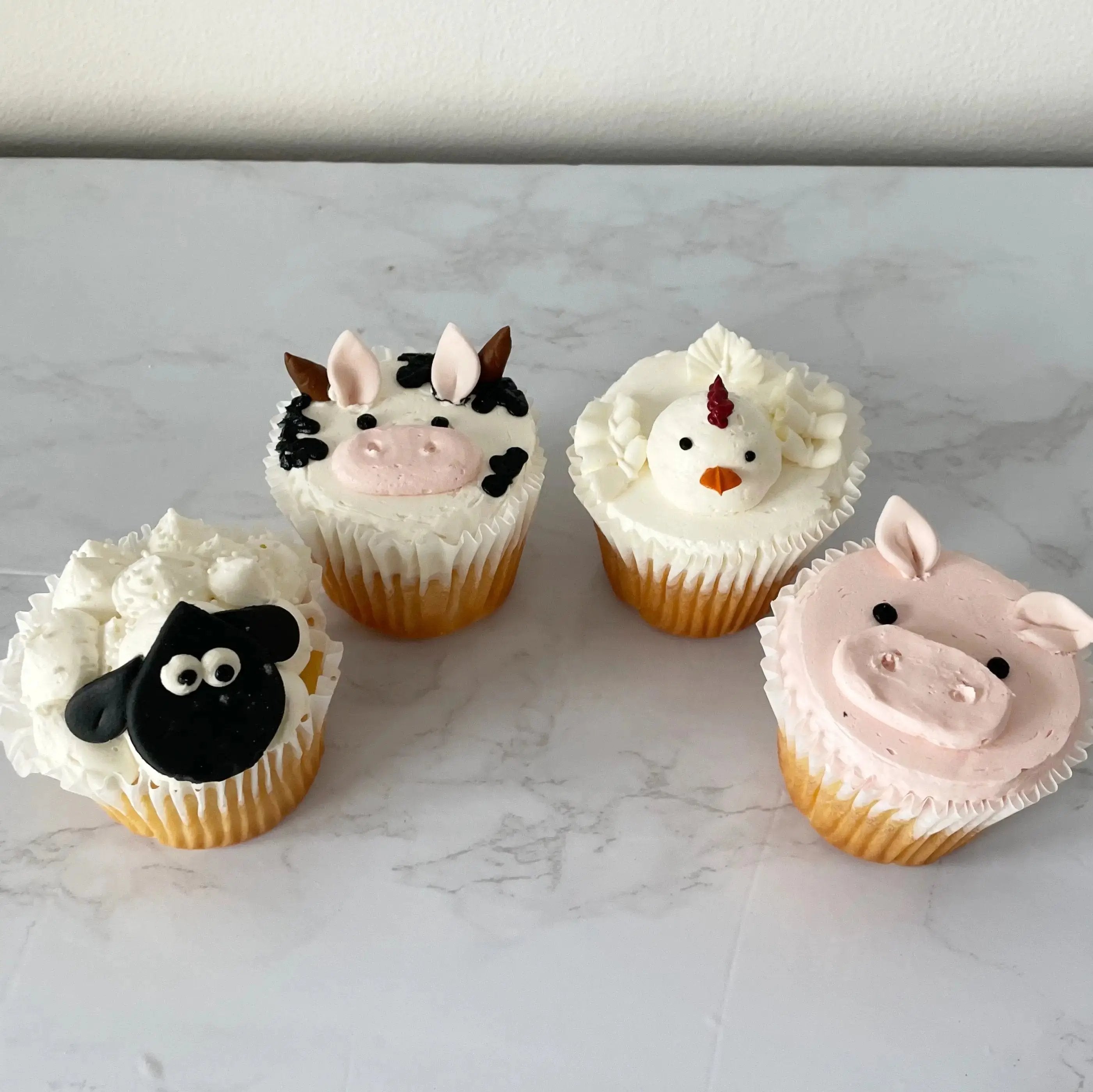 A top-down view of a tray with four cupcakes decorated to look like farm animals: a black-and-white cow, a white sheep, a pink pig, and a white chicken.