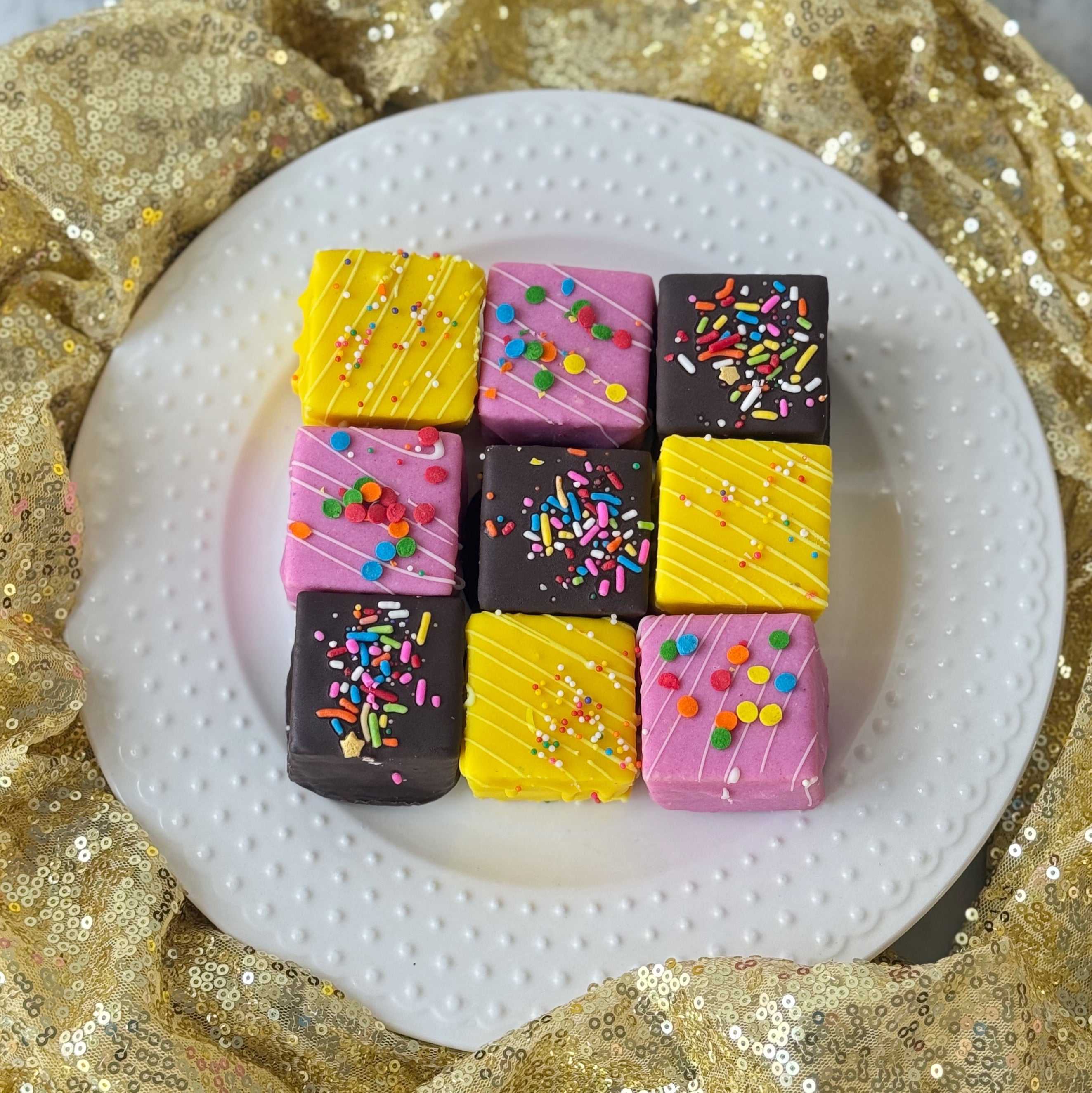 Colorful square petit fours with sprinkles on a white plate against a gold sequin background