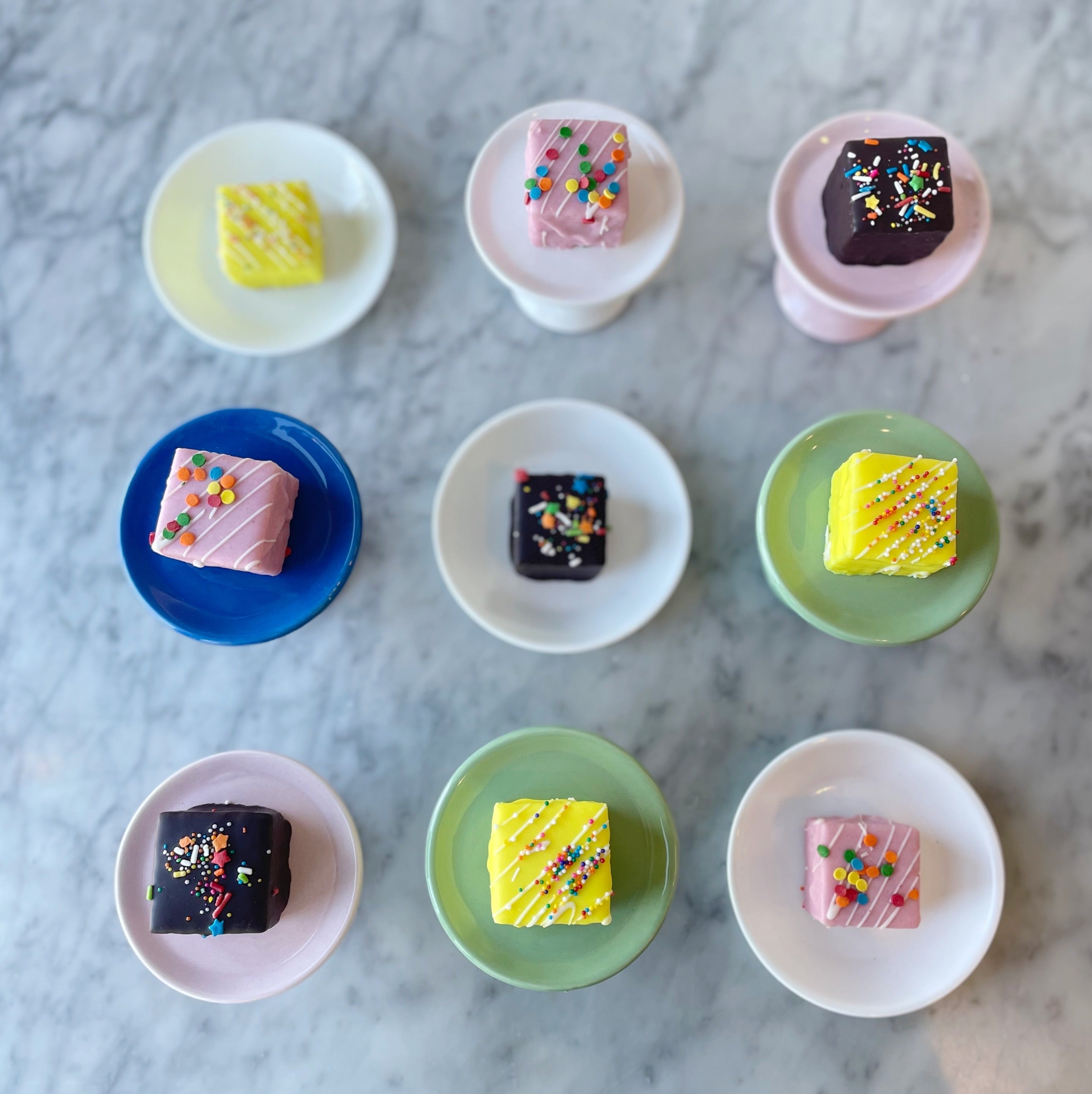 Assorted small petit fours on colorful plates on a marble surface
