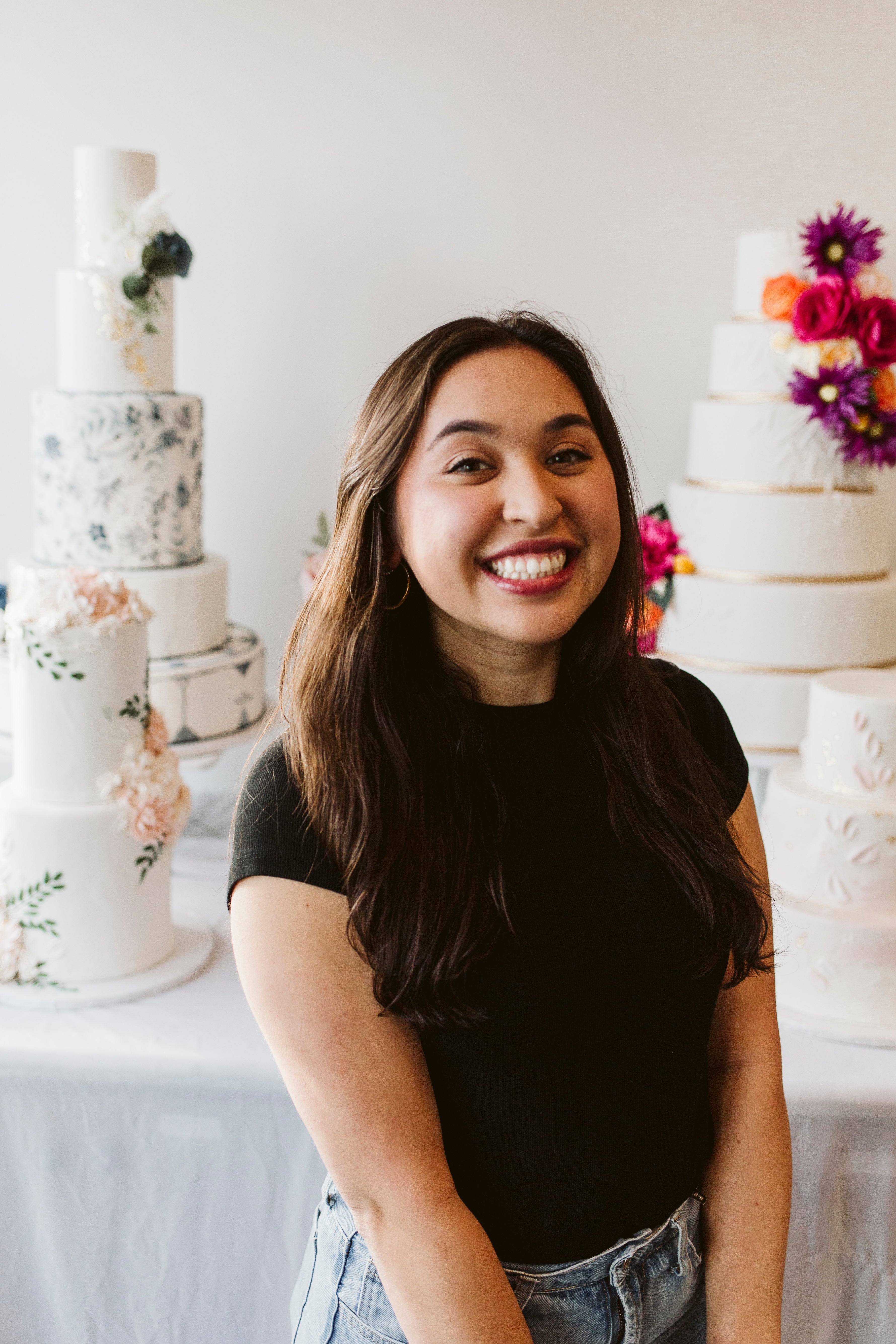 Woman with long brown hair smiling in front of a display of white cakes with floral decorations