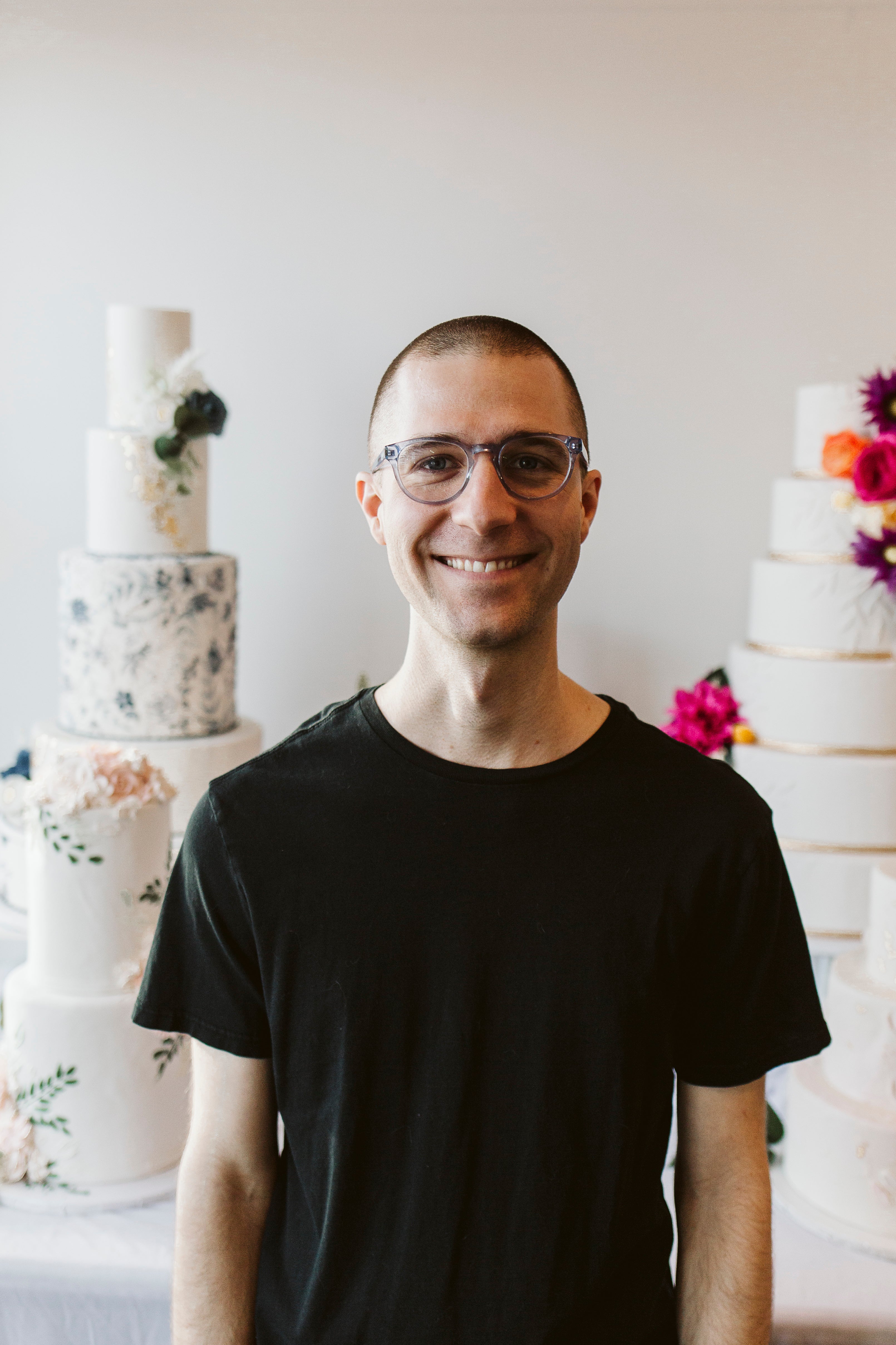 Man in a black t-shirt, smiling in front of a cake display with floral decorations.