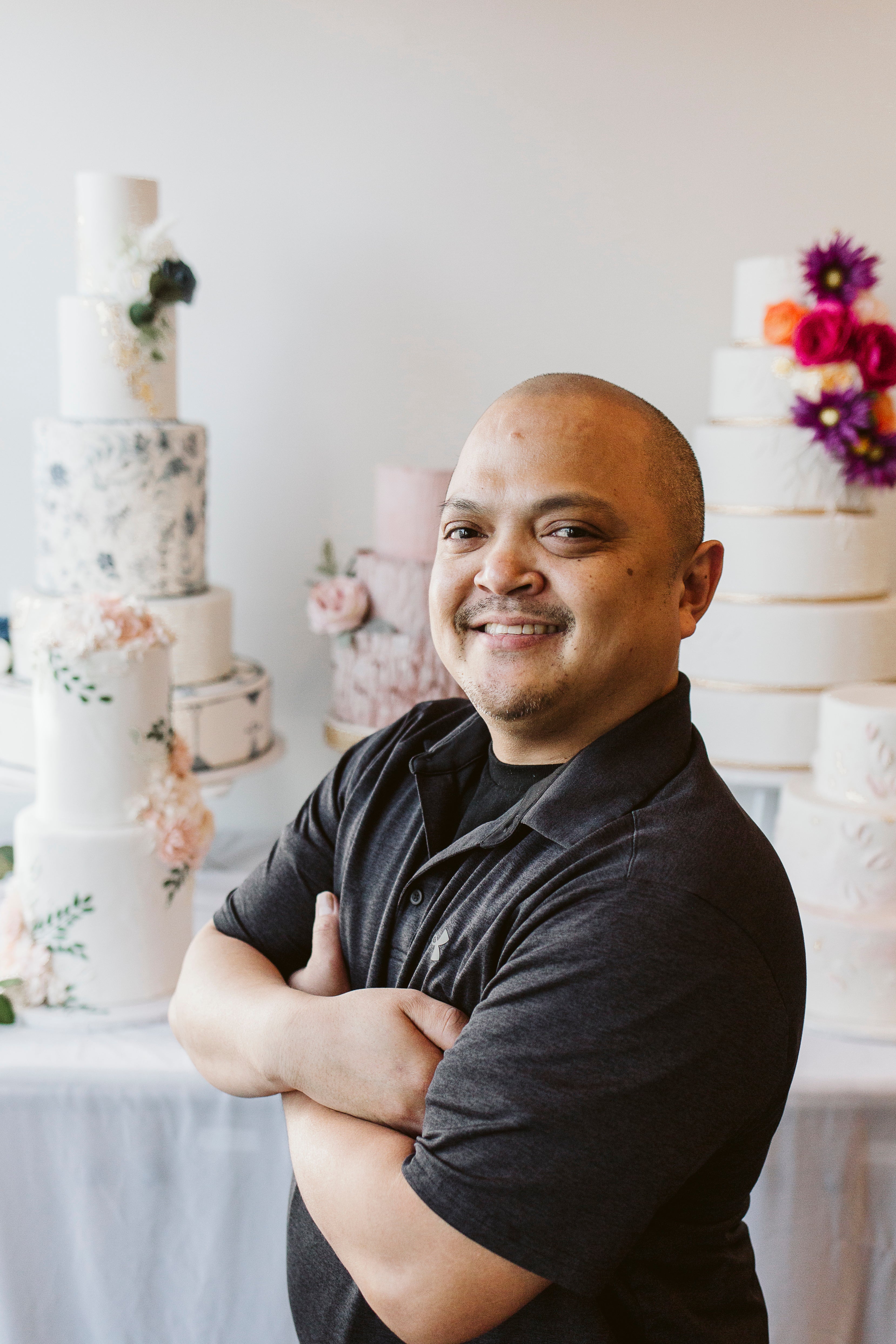 Man with a bald head and beard, smiling confidently while posing in front of decorated wedding cakes