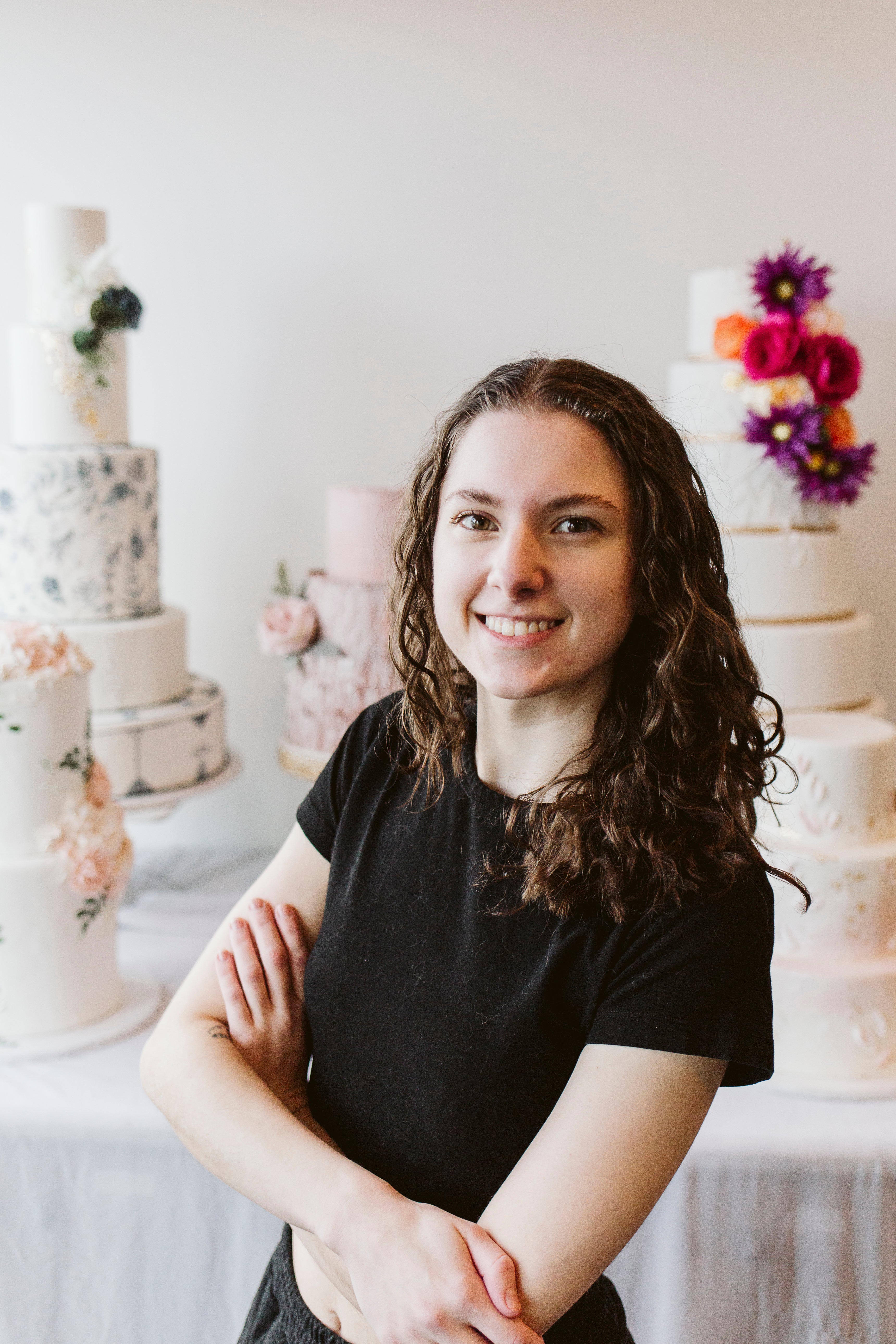 Woman with long dark hair and glasses, smiling in front of cakes adorned with florals in a bright workspace