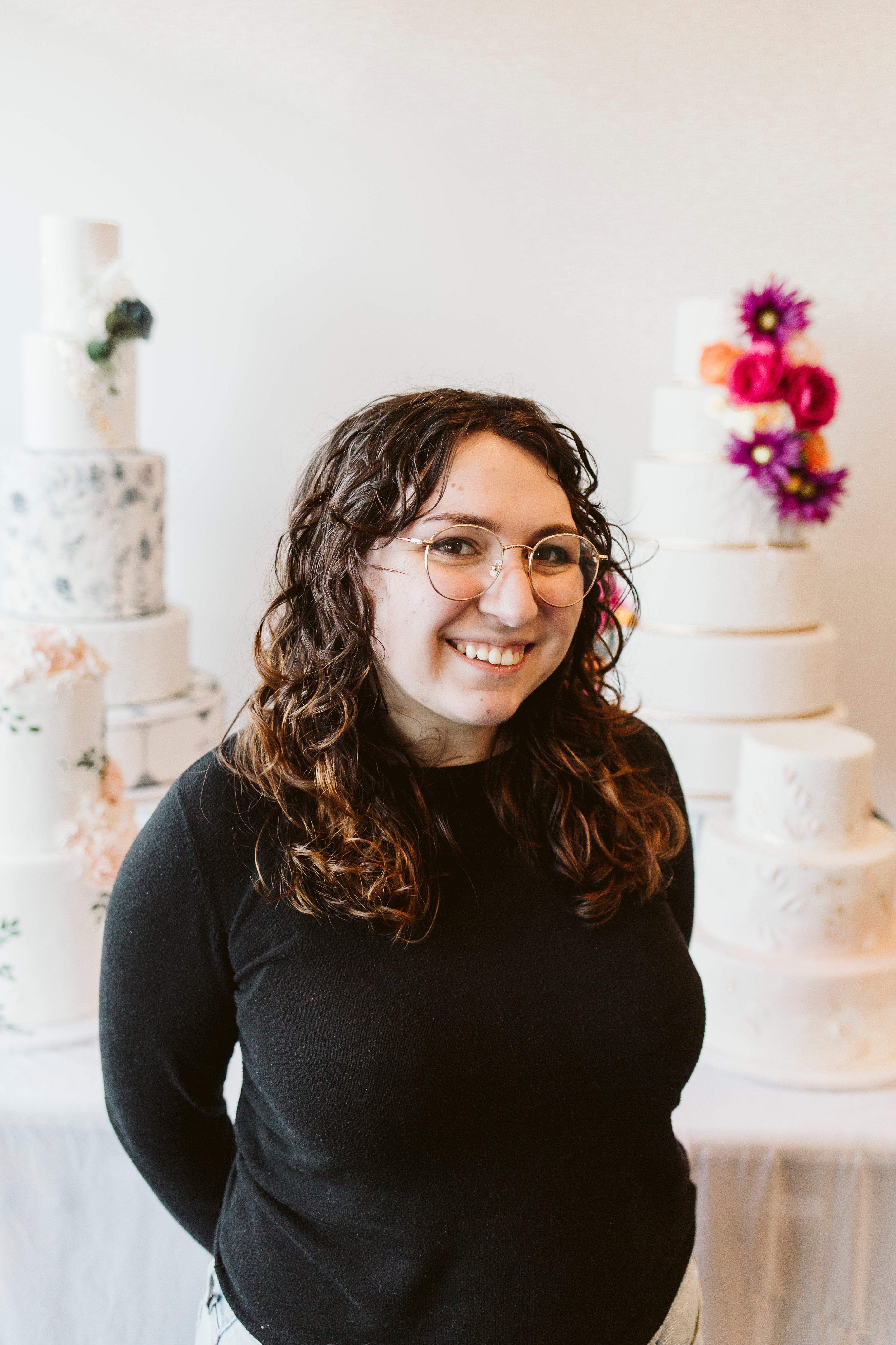 Woman with dark curly hair standing in front of tiered cakes, smiling warmly in a creative studio.