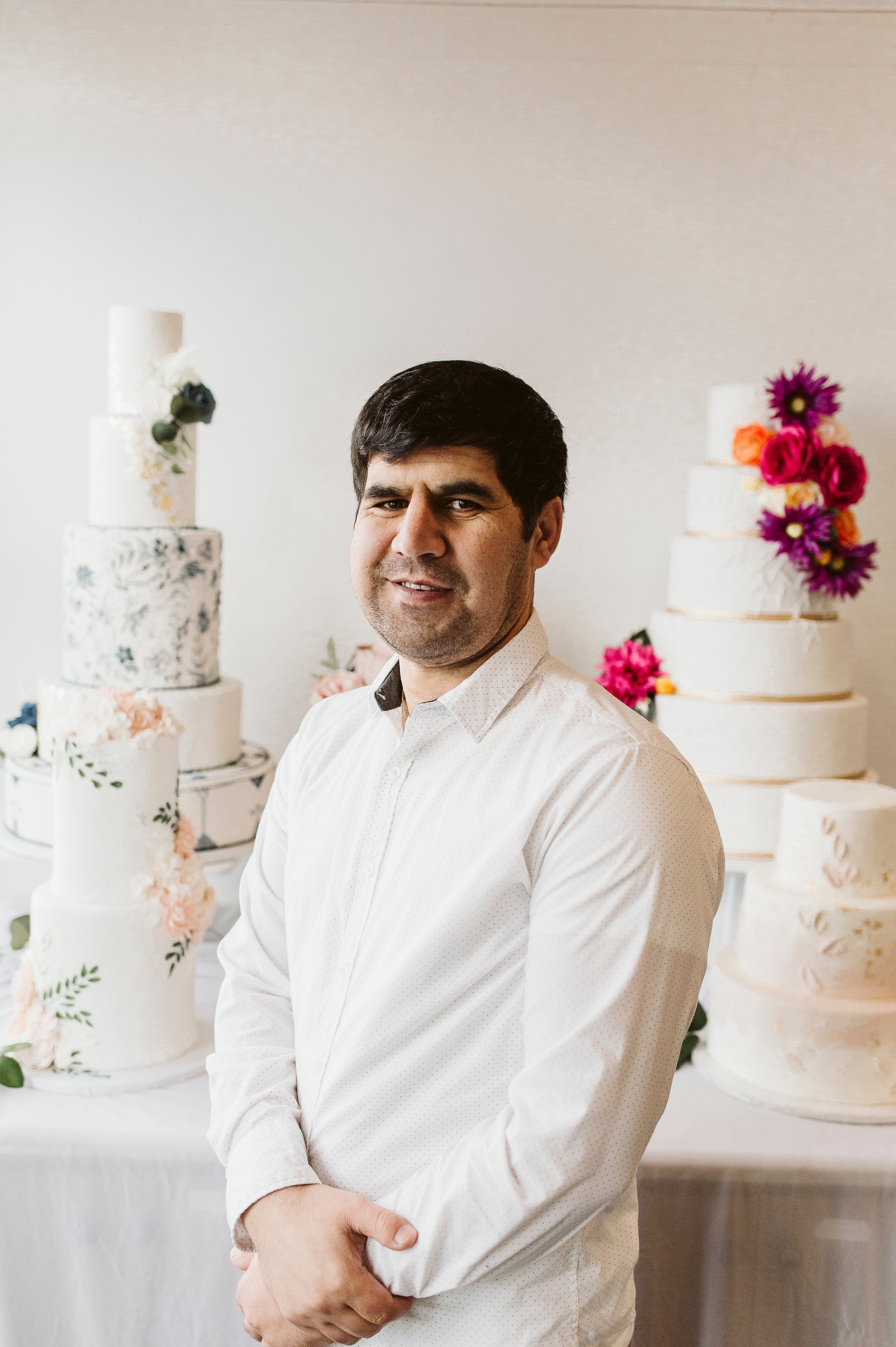 Man in a white chef coat standing in front of tall floral cakes, smiling warmly at the camera