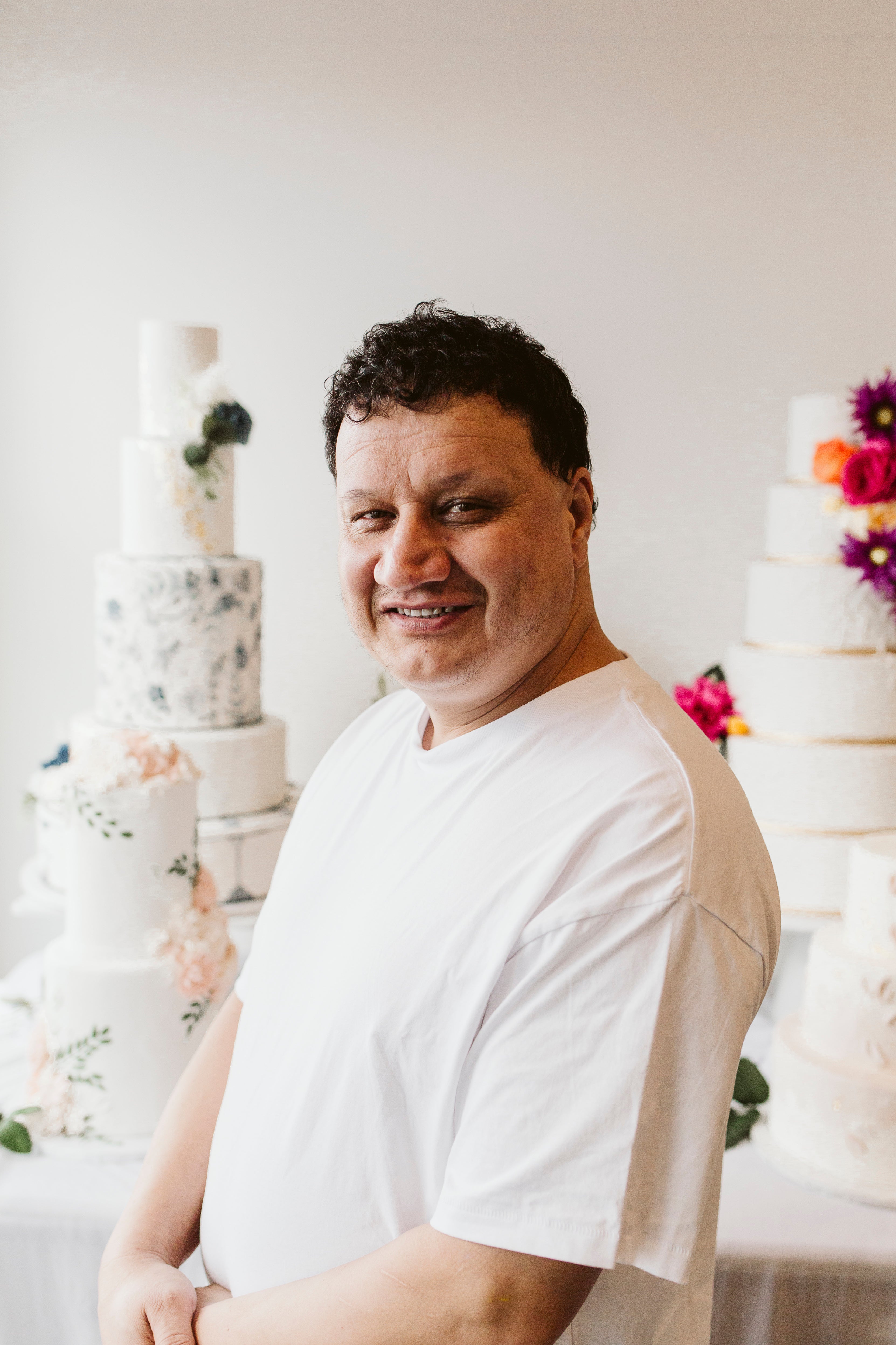 Man in a white chef coat smiling confidently in front of elegant tiered cakes decorated with fresh florals.