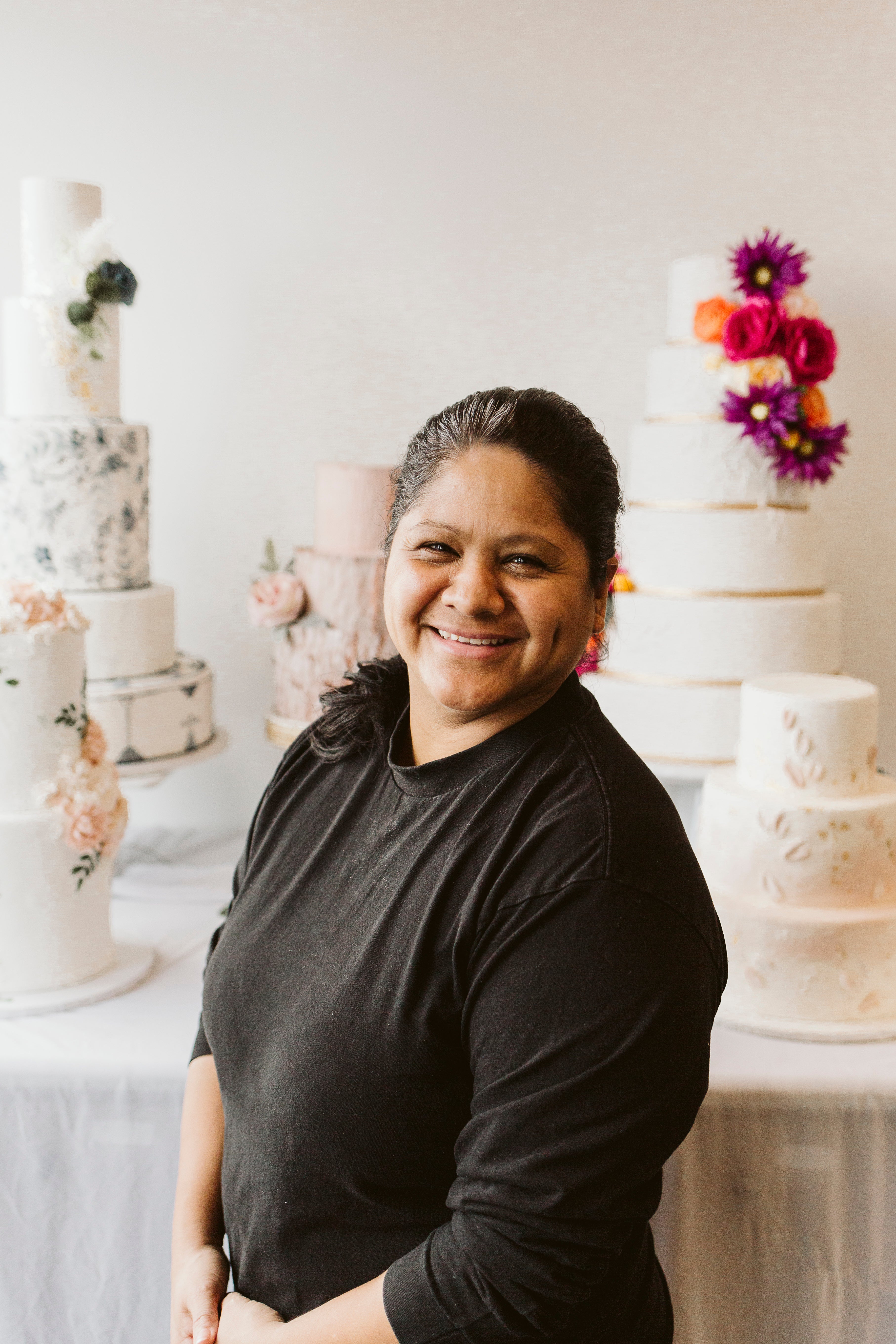 Smiling woman with dark hair and glasses, posing in a bakery with decorated cakes in the background.