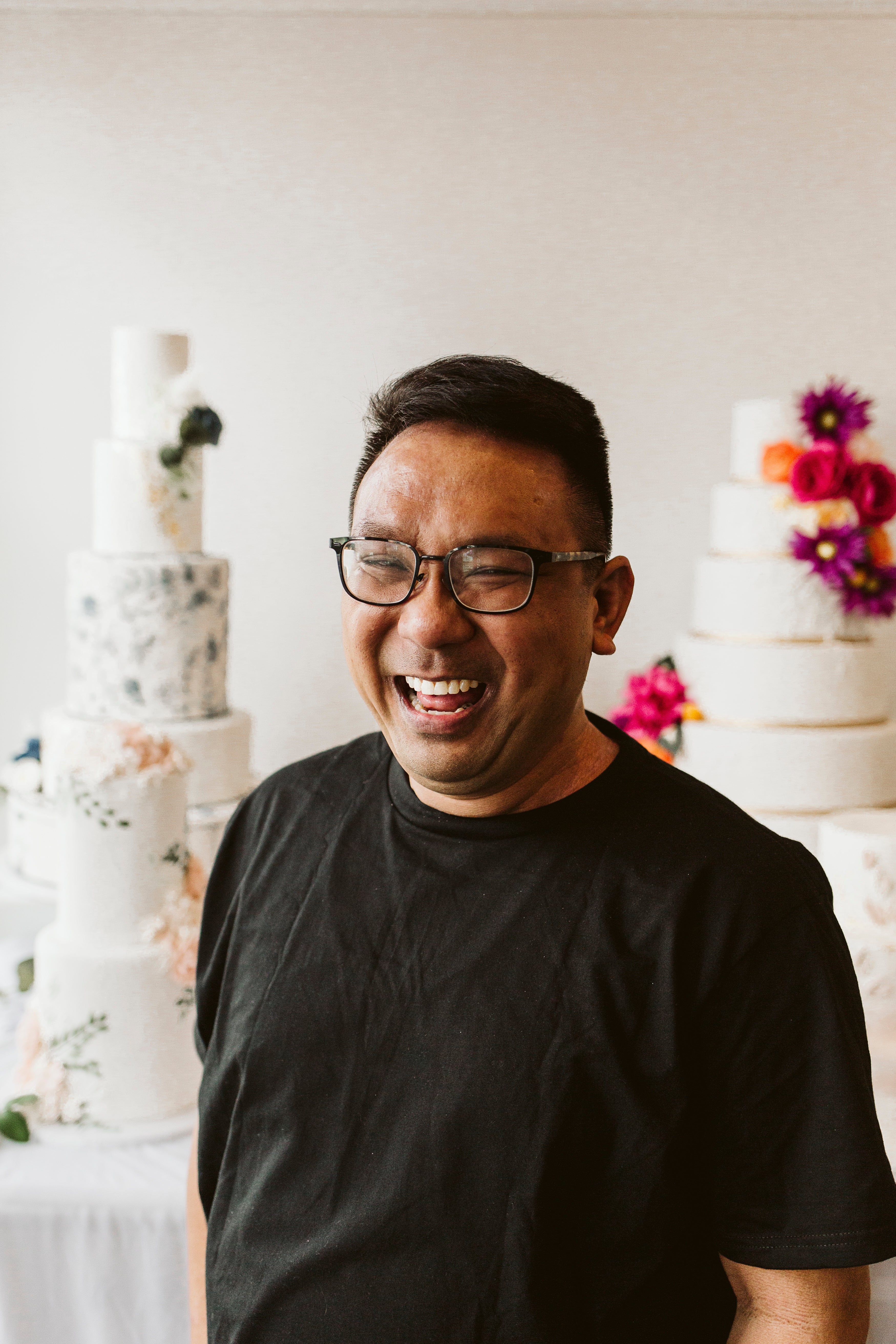 Smiling man in a black shirt standing in front of a display of tall, elegantly decorated cakes with floral accents.