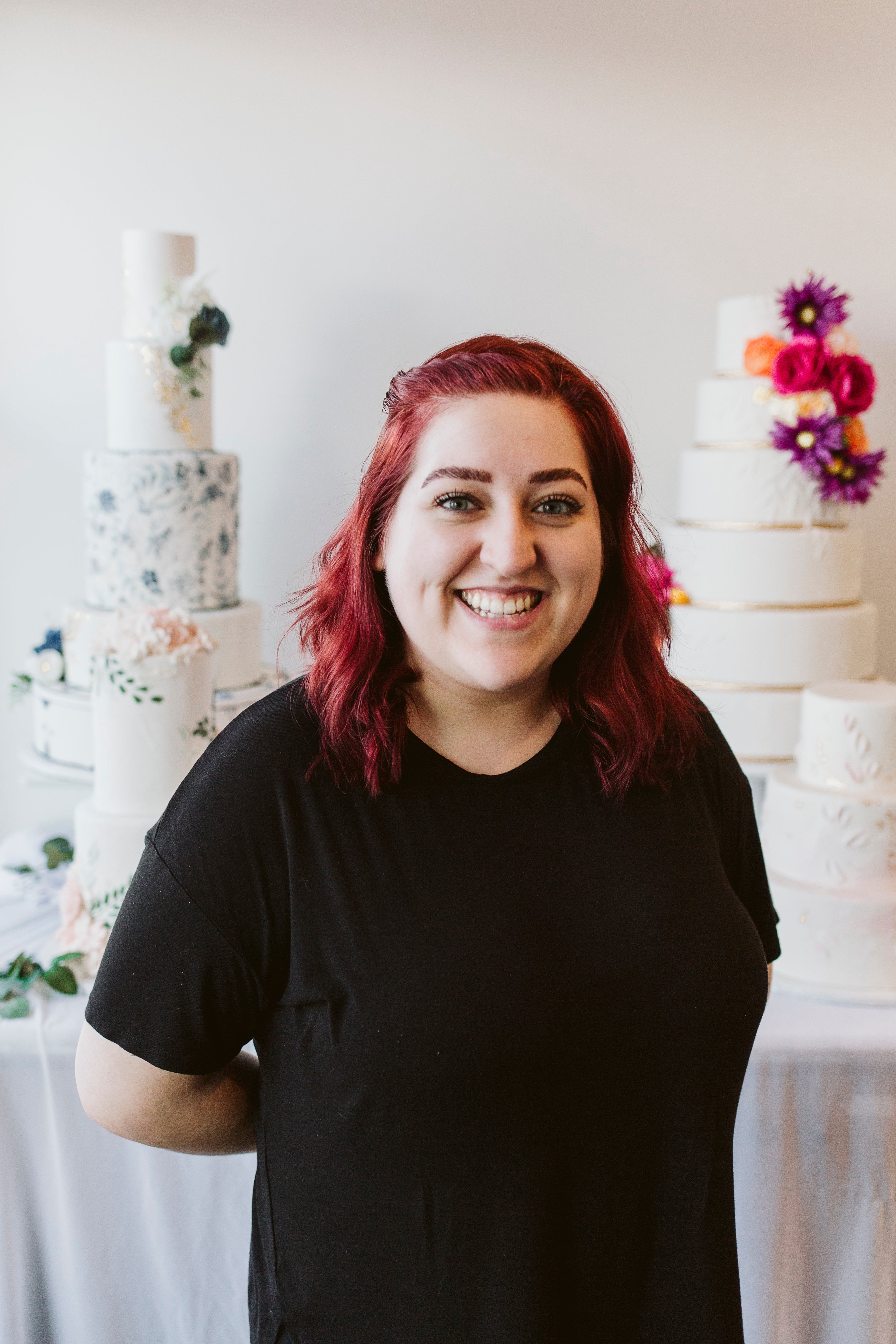 Woman with vibrant red hair wearing a black shirt, smiling in front of white cakes adorned with bright pink flowers