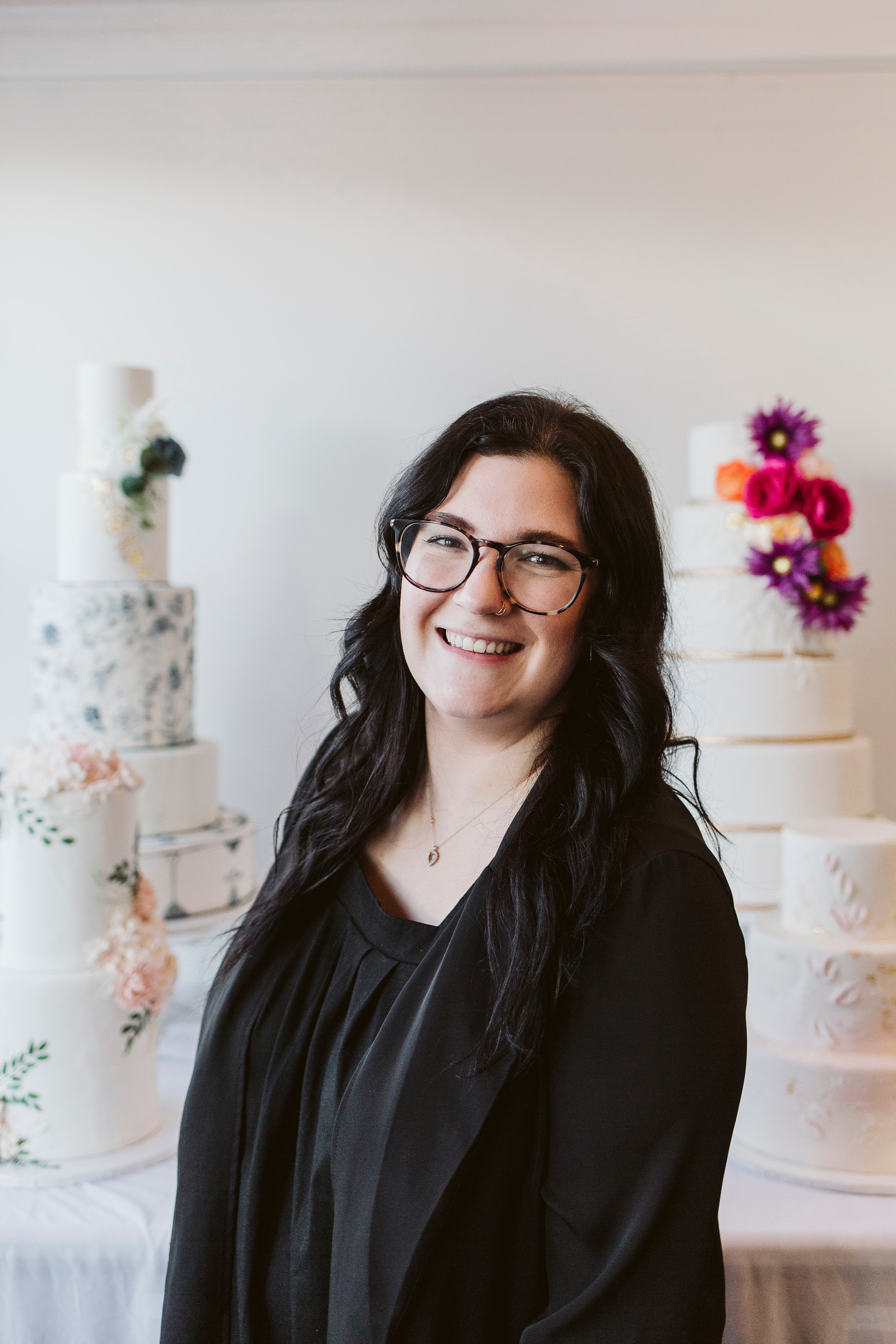 Woman with long dark hair, wearing glasses, smiling in front of decorated cakes in a pastel-toned bakery