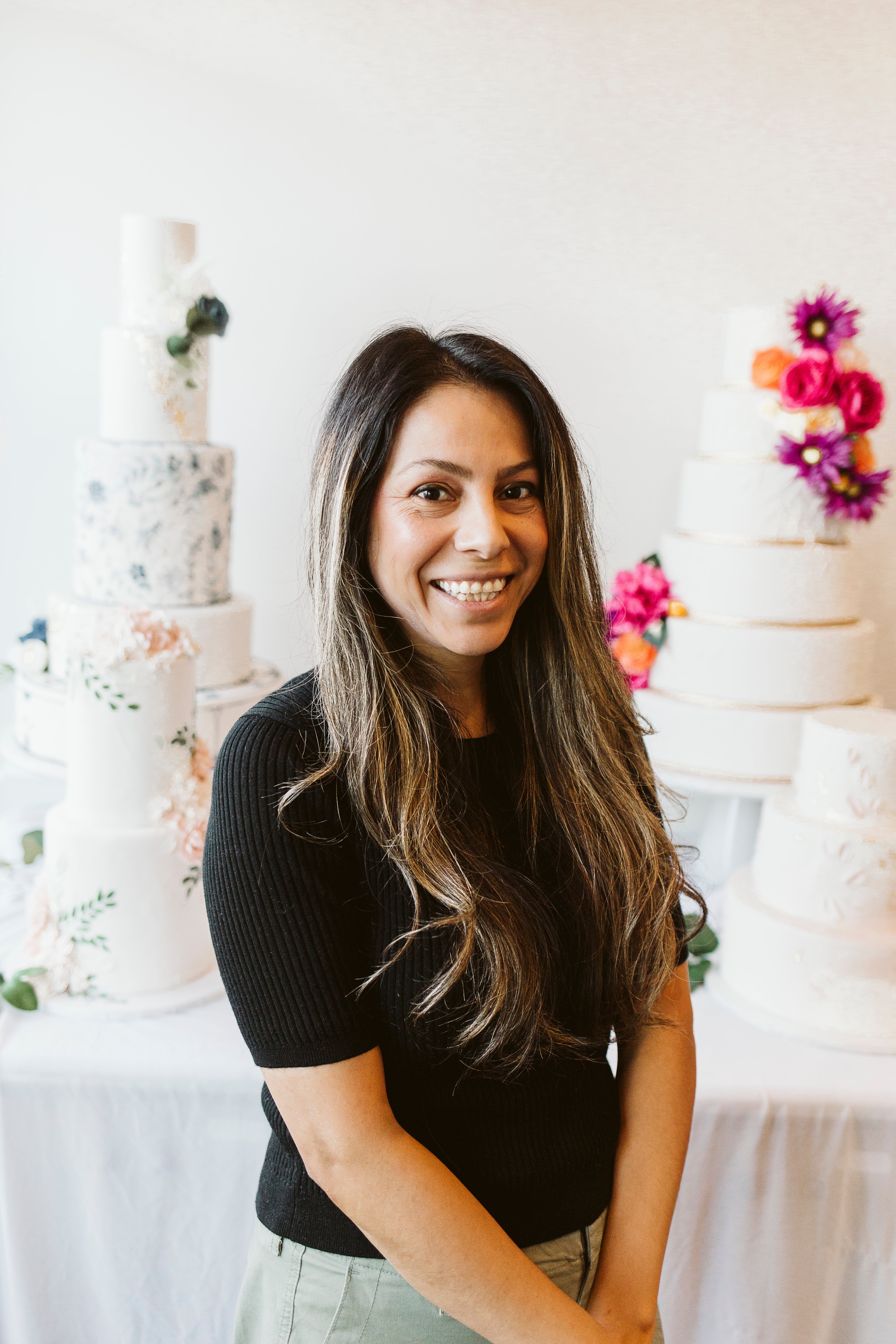 Woman with shoulder-length dark hair posing confidently in a bakery setting with pink and white cakes behind her.