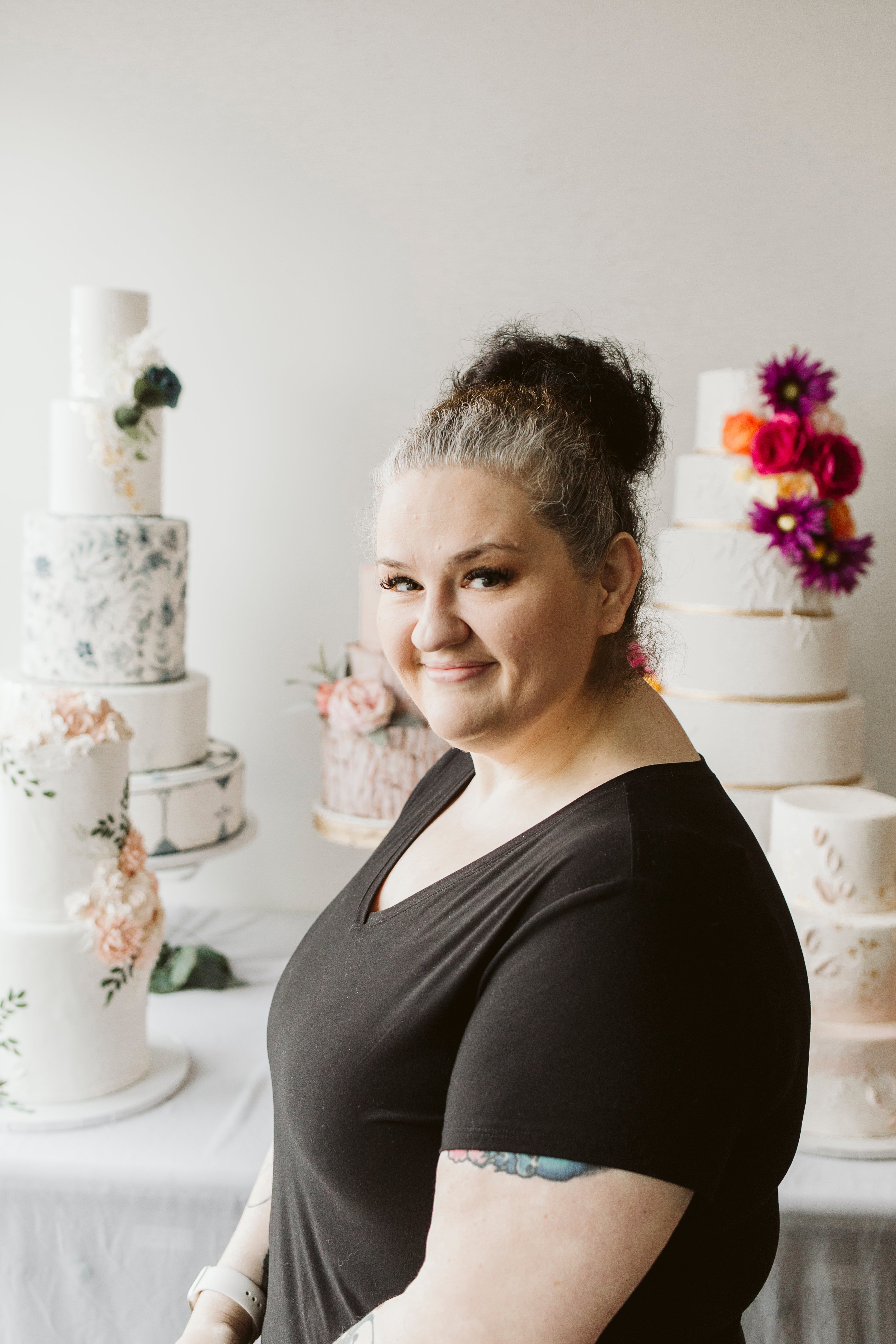 Woman in a black top with red lipstick and a red flower in her hair, smiling in a bakery setting.
