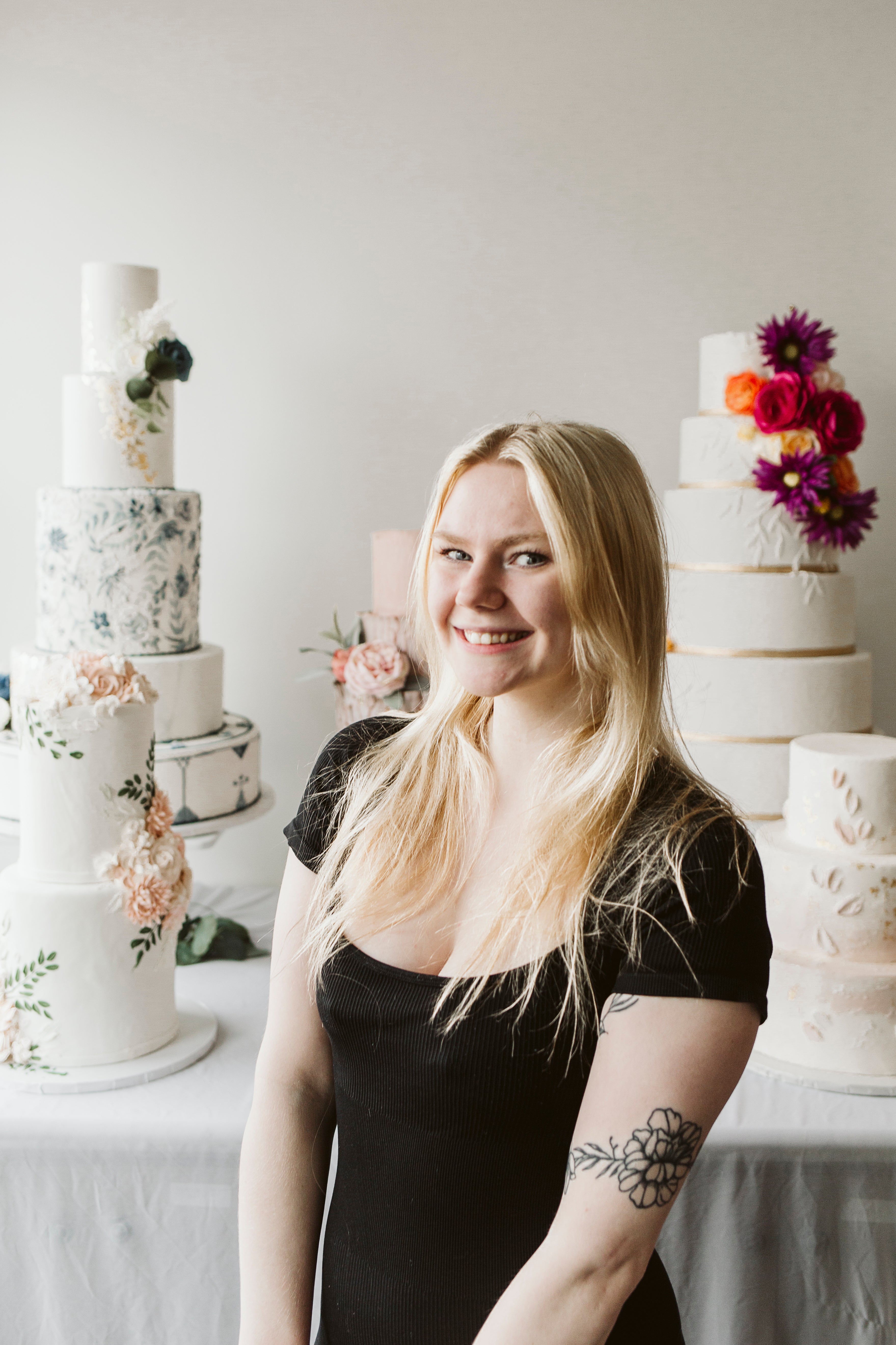 Portrait of a woman in a black dress with blonde hair, smiling in front of an ornate white cakes.