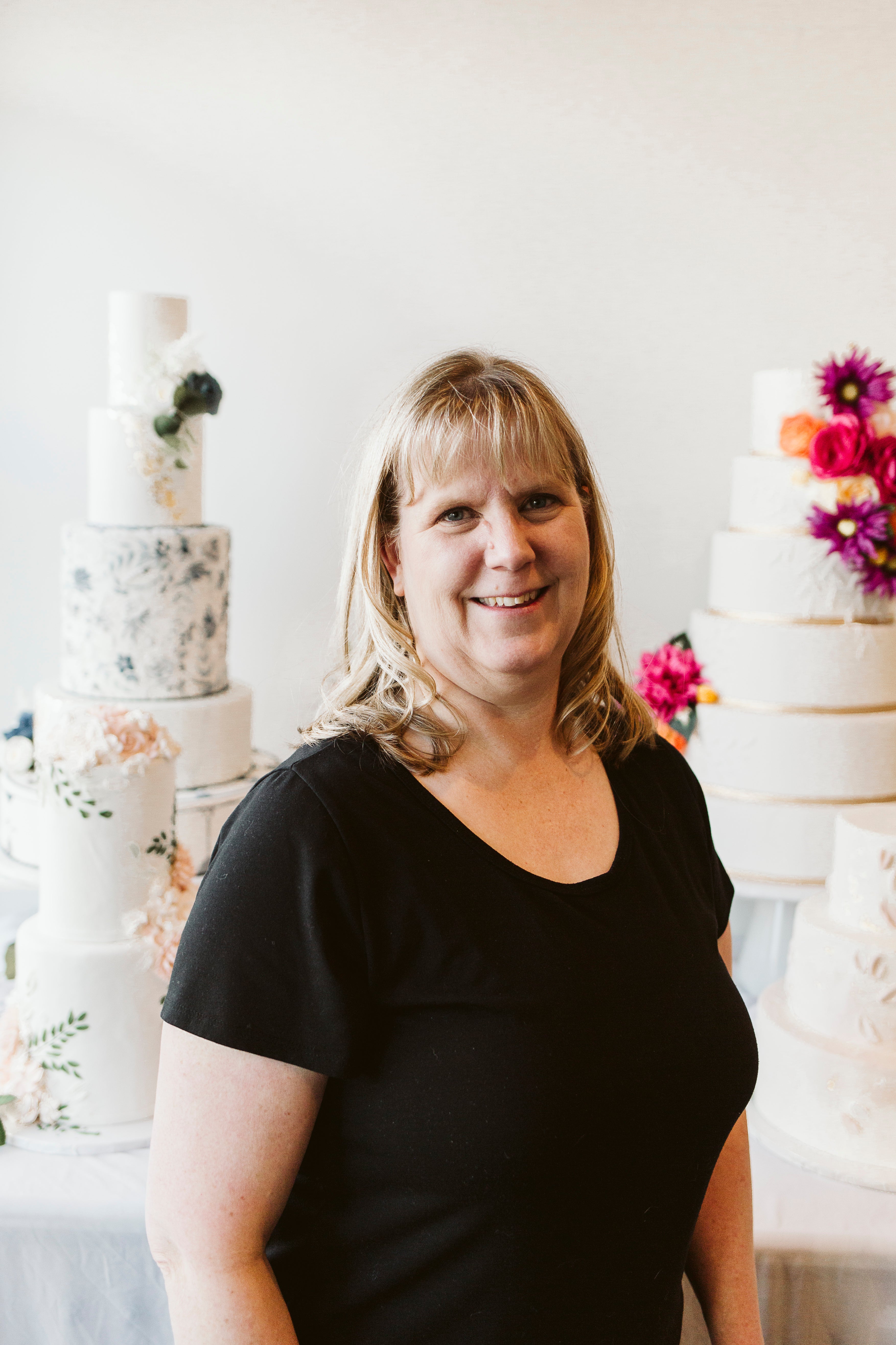 Woman with blond hair and  smiling in a black shirt, standing in front of white cakes with pink flowers