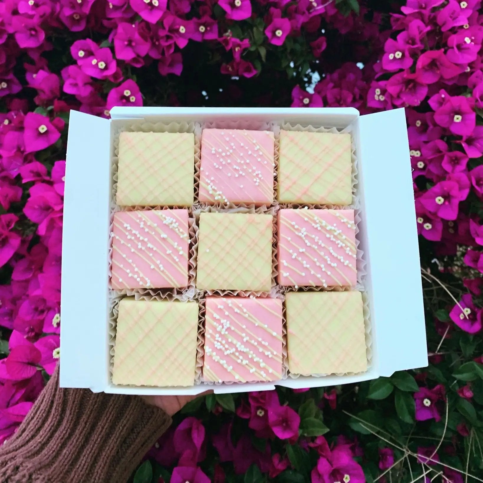 Box of nine pastel-colored petit fours in pink and yellow, displayed against vibrant pink bougainvillea flowers
