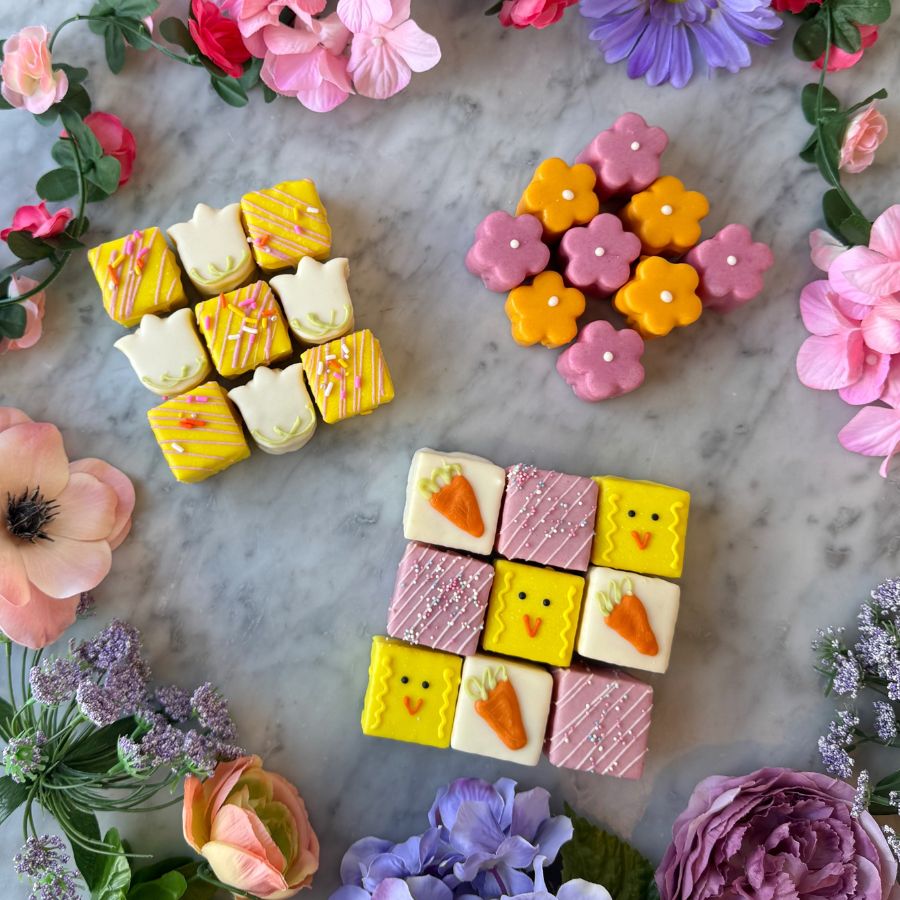 Colorful petit fours that are square-shaped and Easter-themed items on a marble surface with flowers around.