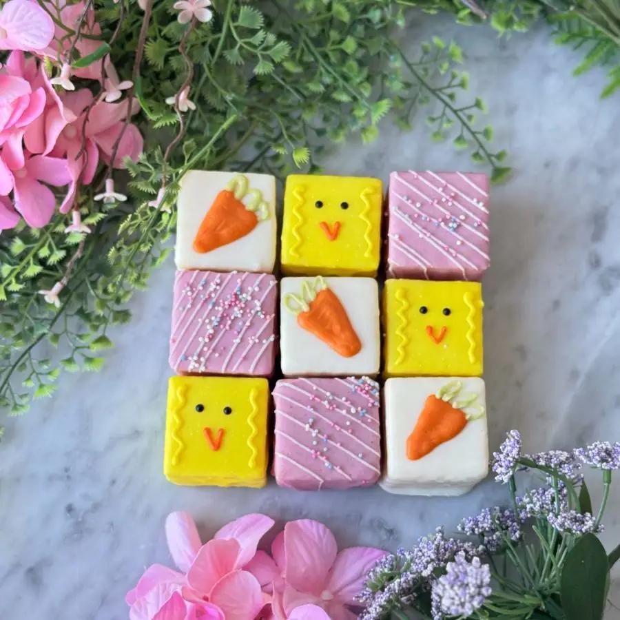 Decorative petit fours that are square-shaped with a carrots and chicks piped design on a marble surface with flowers.