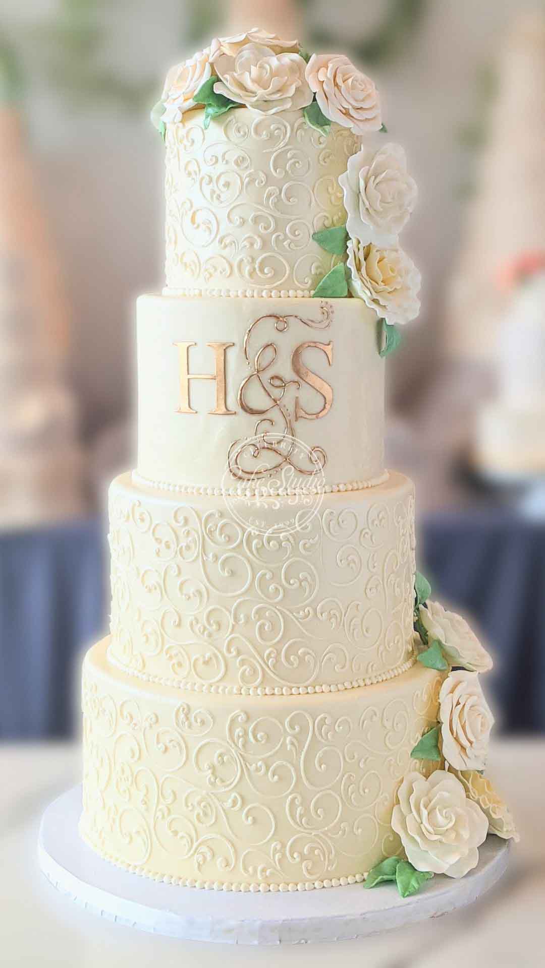 A tall, multi-tiered white wedding cake with textured ruffles on the bottom two tiers and gold lettering on the top tier. The cake is decorated with a cascade of pinkish white flowers.