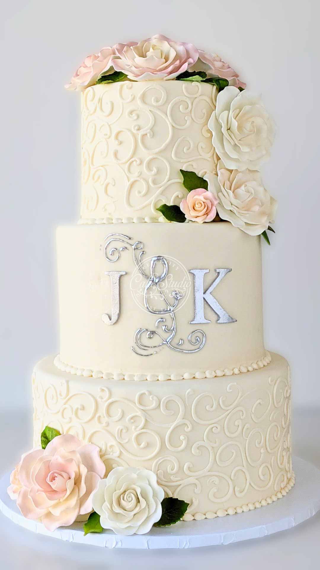 A three-tiered white wedding cake decorated with white and pink flowers. The second tier has a large "J & K" monogram in silver.