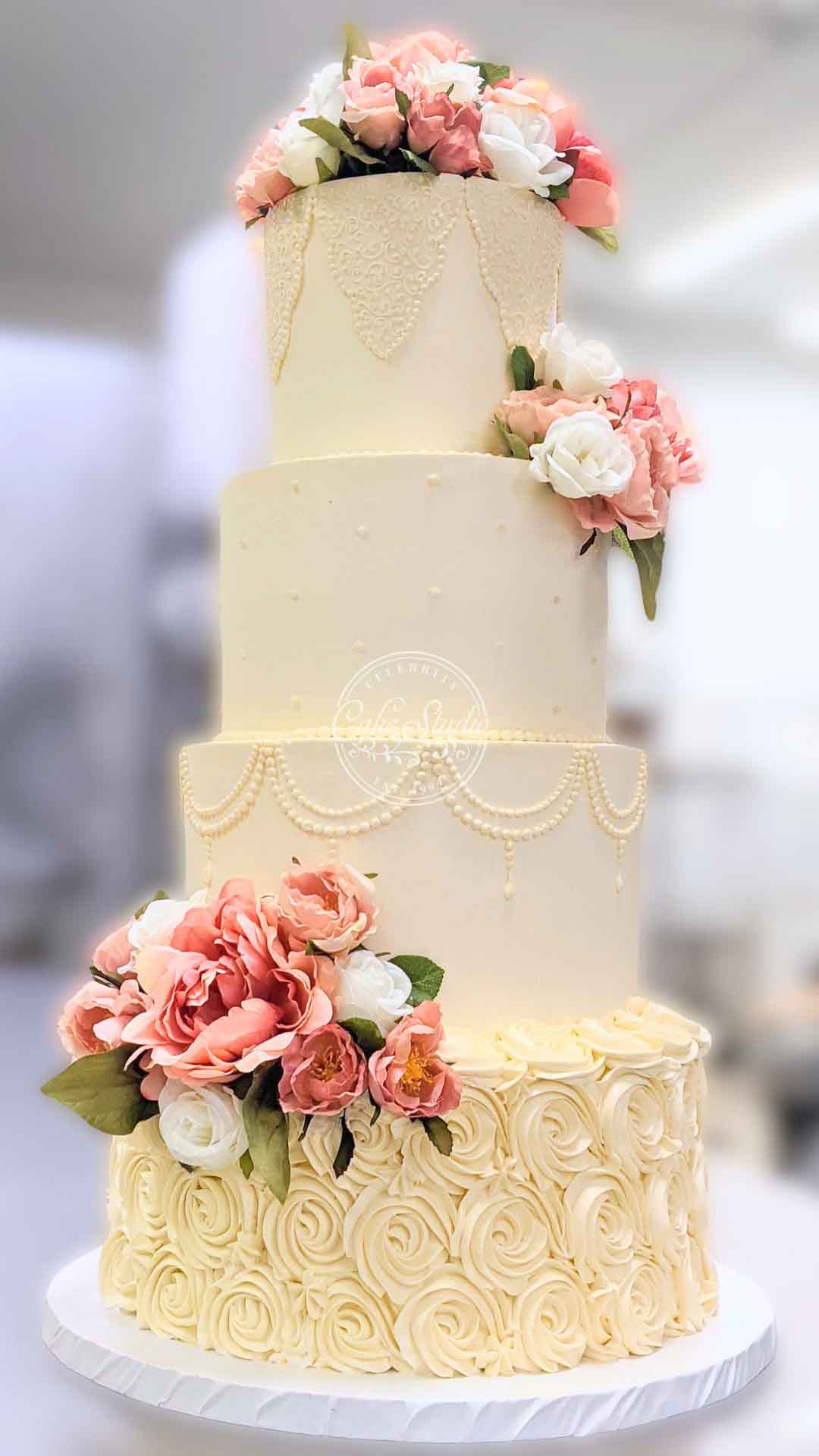 A four-tiered white wedding cake. The bottom tier is ruffled, and the cake is decorated with pink and white flowers cascading down the side.
