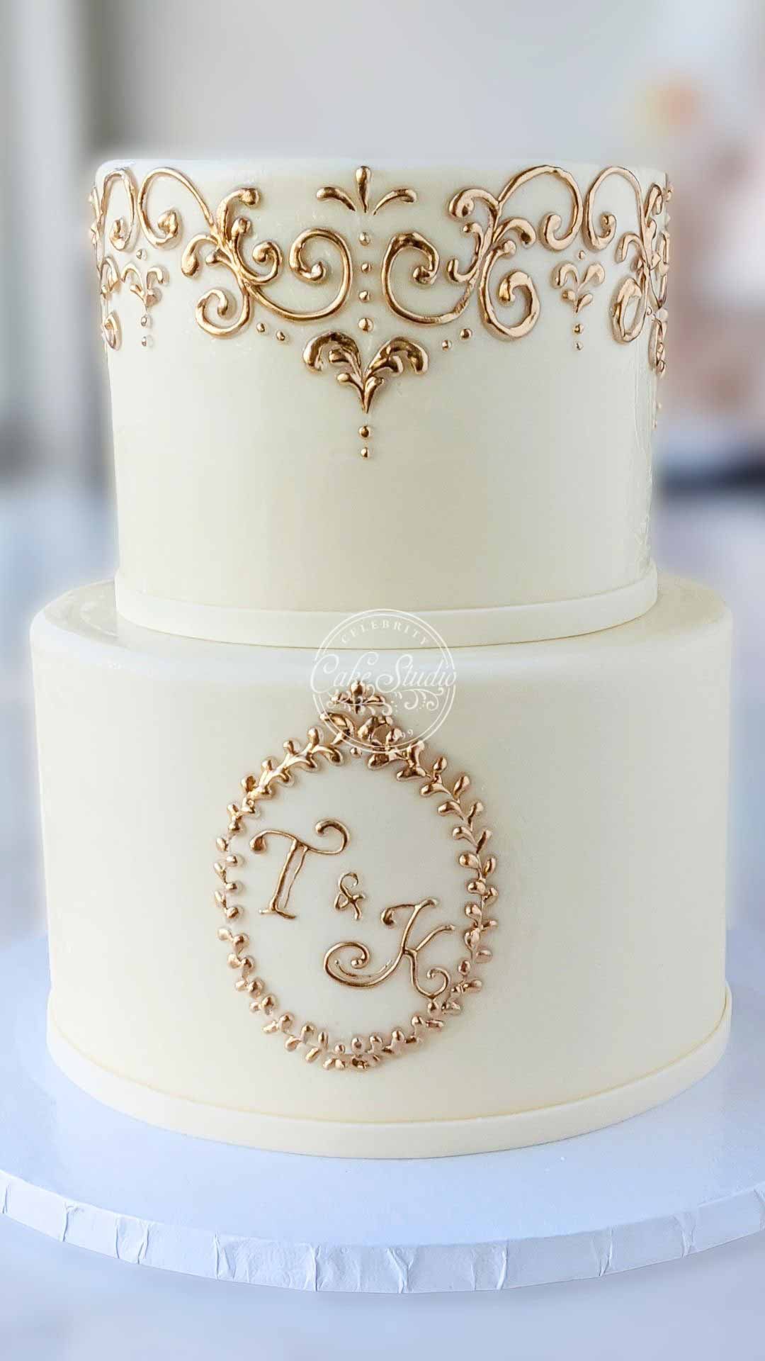A two-tiered white cake with an ornate gold design on the top tier and a gold, framed monogram on the bottom tier.