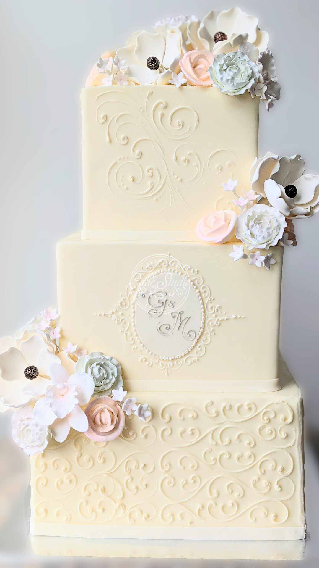 A three-tiered white square wedding cake with a textured pattern. It's decorated with pink, white, and blue flowers trailing down the side.