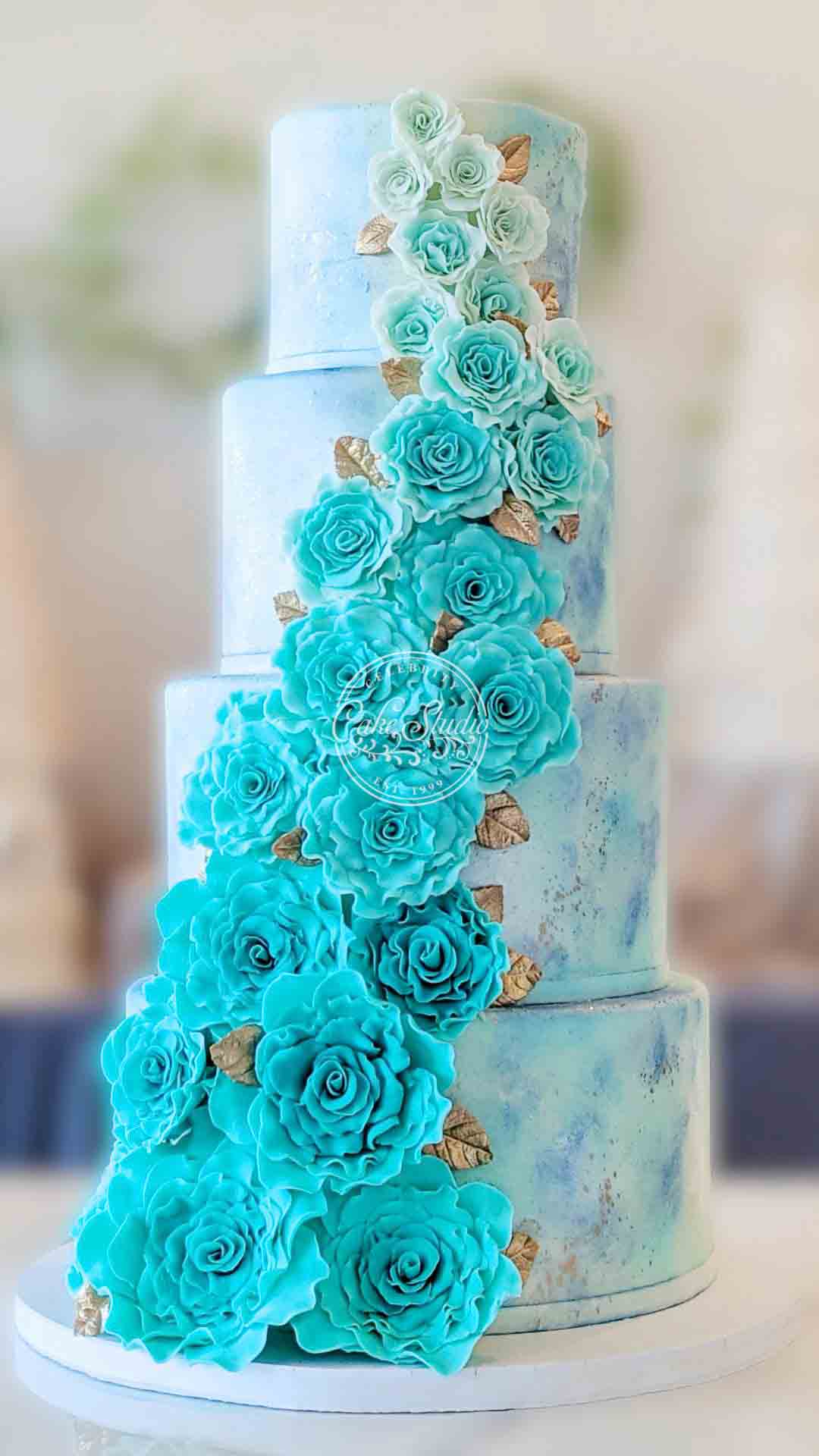 A tall, multi-tiered white and light blue marbled cake with a cascade of dark turquoise roses down the side.