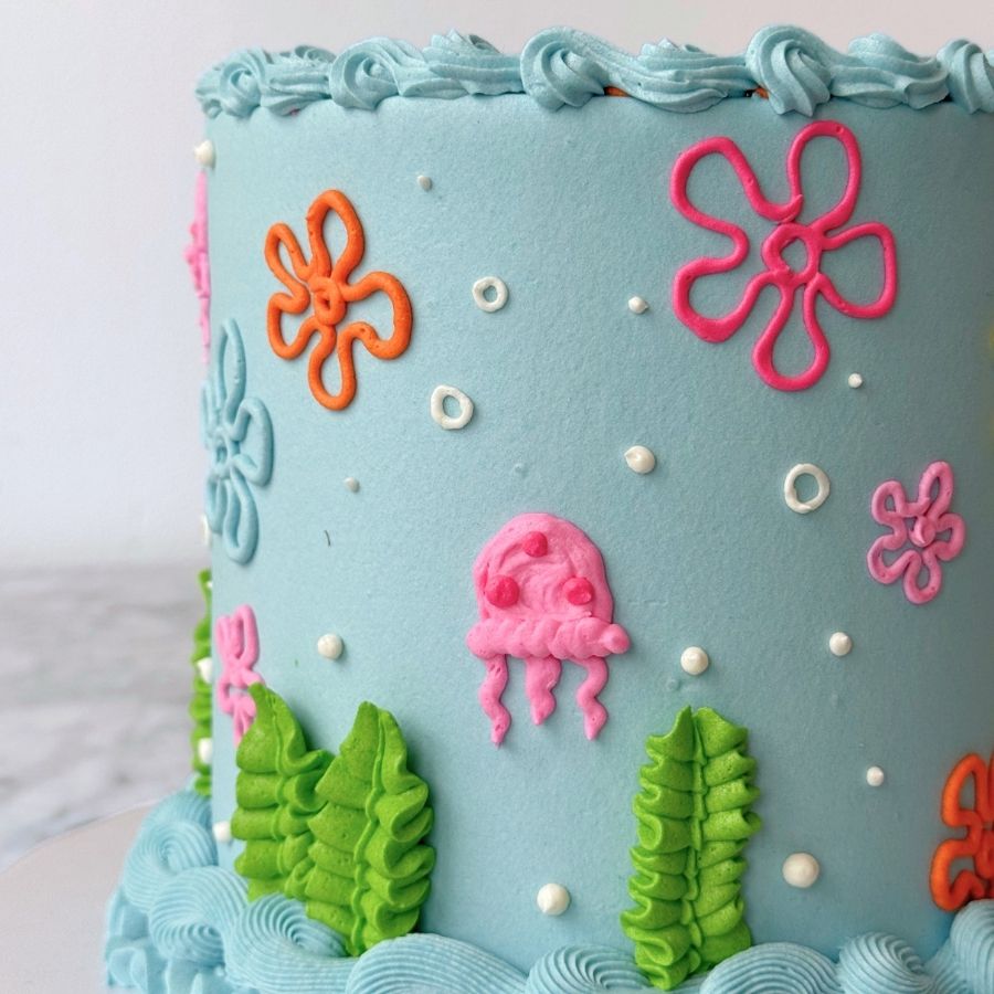 A detailed close-up of the side of a blue cake. It features a hand-piped pink jellyfish, bright green seaweed at the base, and various colorful floral patterns. Small white sugar pearls are scattered across the surface to represent bubbles.