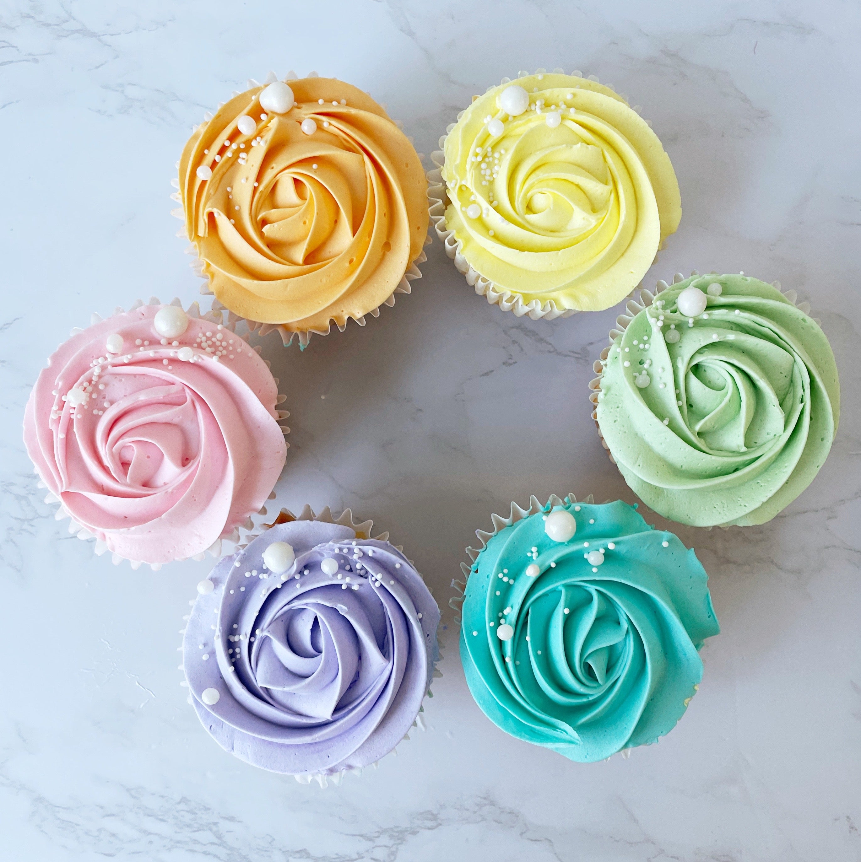 A top-down view of six cupcakes with buttercream roses in pastel rainbow colors arranged in a circle