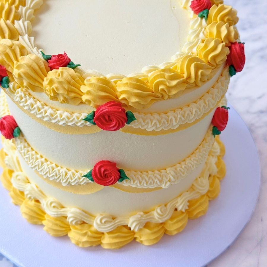 A close-up side view of a vibrant yellow tiered cake. The design features elaborate buttercream swags, golden sugar pearls, and a large red rose decoration with green leaves. The piping includes vintage-style ruffles and shell borders.