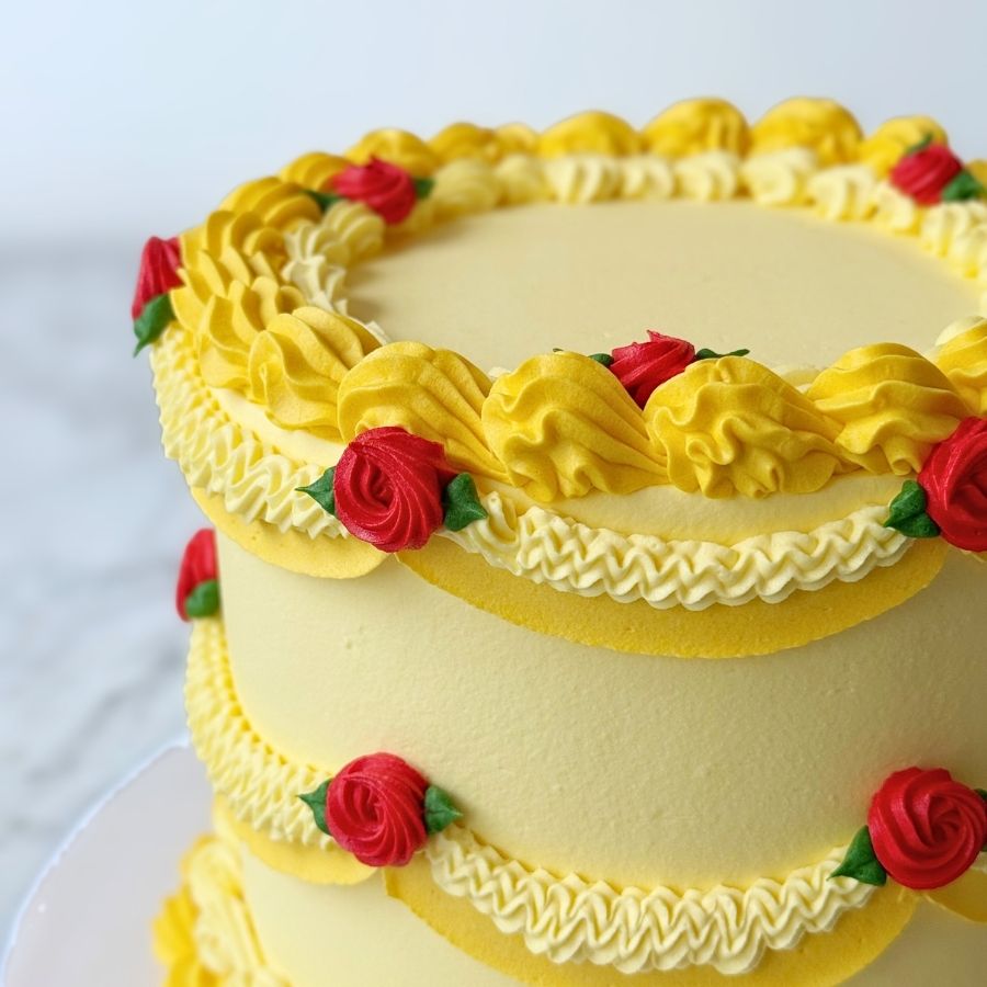 An overhead view of a round yellow cake. The top is smooth and surrounded by a thick, ornate border of yellow buttercream ruffles. The rim is decorated with shimmering gold beads and small hand-piped red roses.