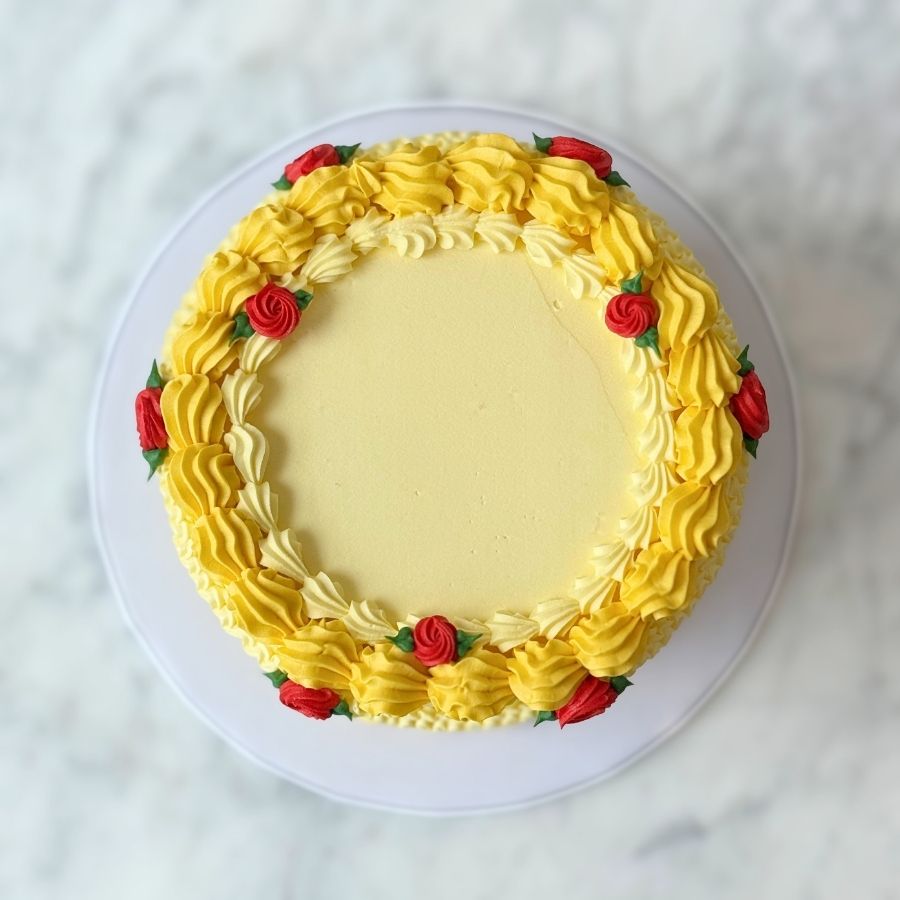 A macro detail shot of the side of the cake, highlighting the intricate yellow buttercream piping. The image shows symmetrical swags and vertical ruffles accented by gold sugar pearls and a small, delicate red rose blossom.