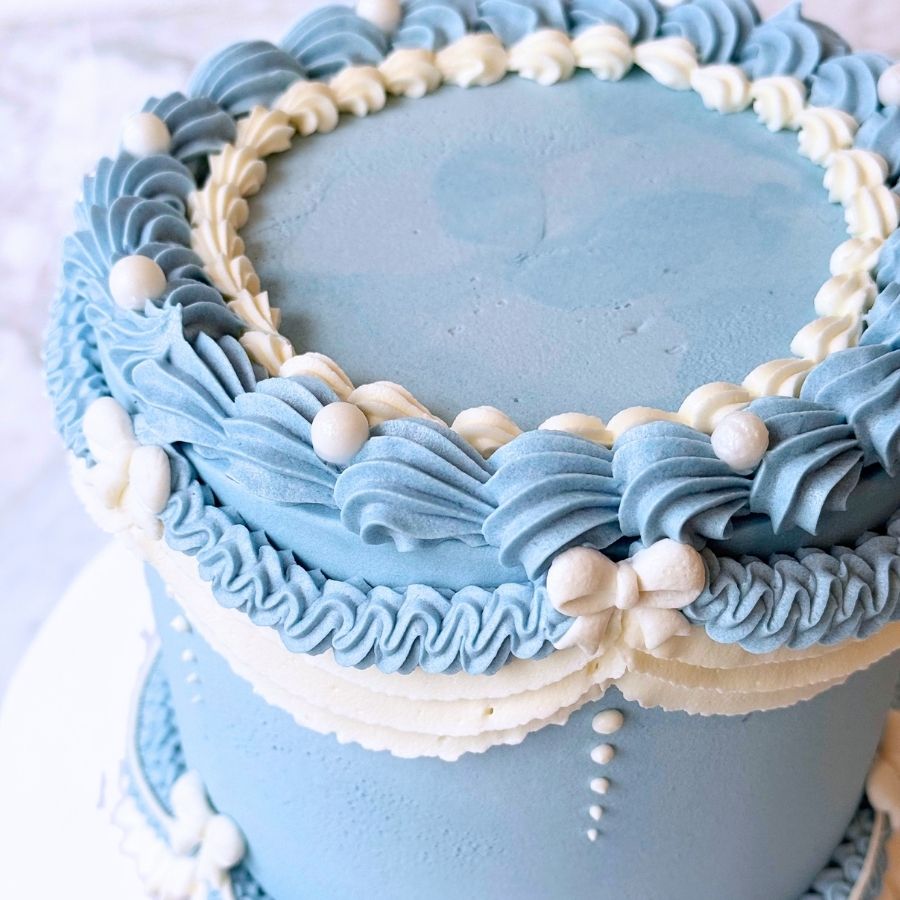 An overhead view of a round light blue cake. The top is smooth and surrounded by a thick border of light blue and white frosting swirls. It is decorated with small pink flowers and gold beads.