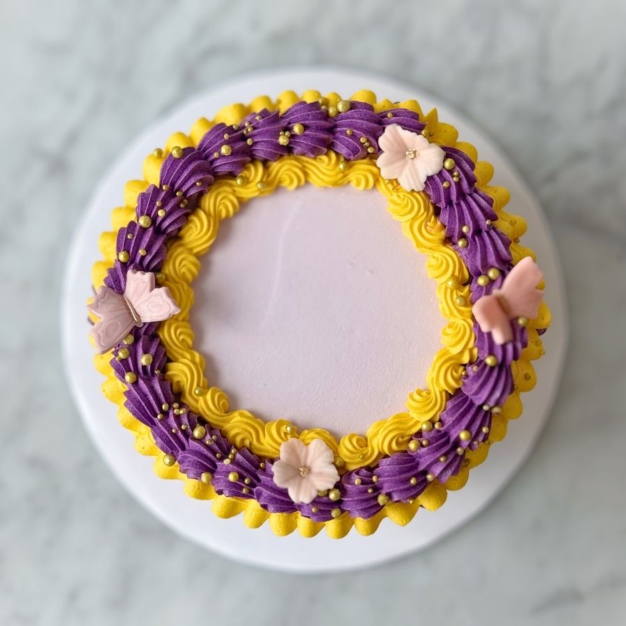 An overhead view of a round cake with smooth lavender frosting on top. The outer edge is decorated with a thick, ornate crown of purple and yellow buttercream ruffles, featuring gold pearls, small pink blossoms, and sugar butterflies.