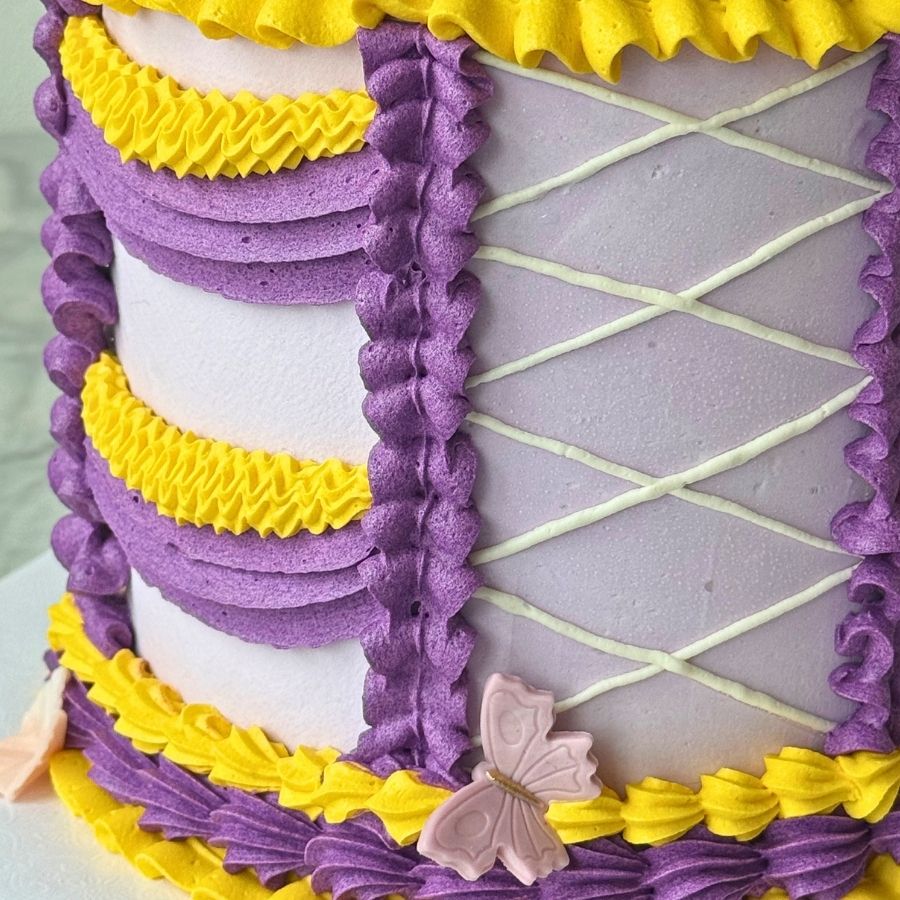 A macro shot focusing on the side of a themed cake. The design includes a white piped "corset" lace pattern on a lavender background, framed by vertical purple ruffles and horizontal yellow buttercream swags. A small pink butterfly sits near the bottom border.