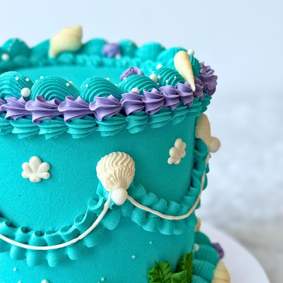 An overhead shot of a round mermaid cake. The top surface is a smooth teal buttercream, encircled by an ornate border of purple and teal ruffles. The rim is embellished with gold beads, delicate pink blossoms, and white starfish and seashell sugar decorations.