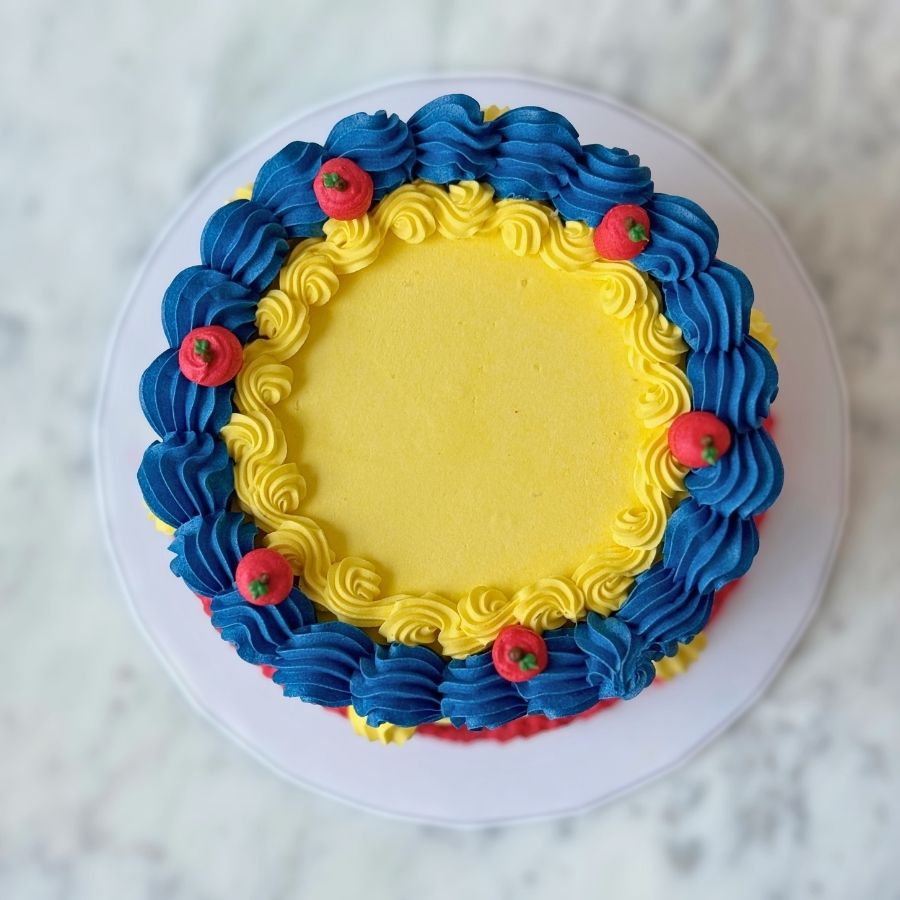 An overhead view of a round dark blue cake. The top is smooth and surrounded by a thick border of dark blue and red frosting ruffles. It is decorated with small gold beads.