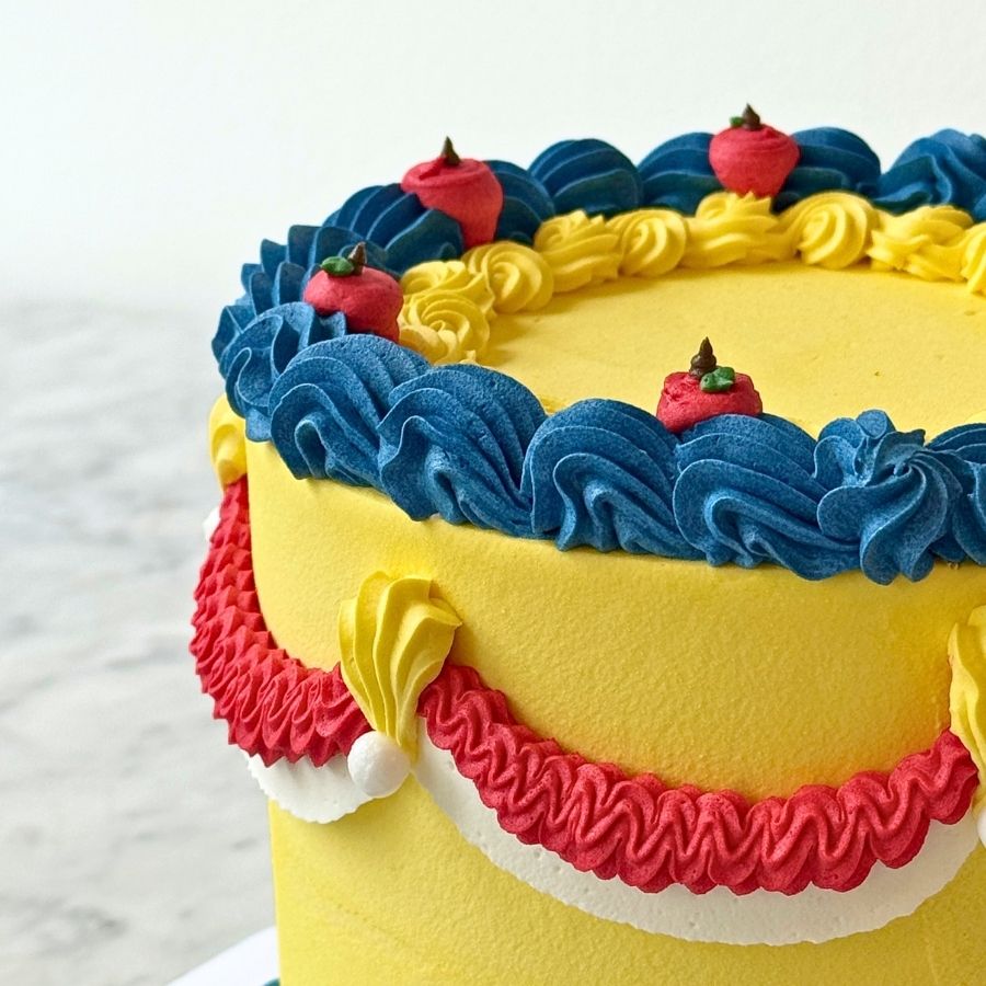 A close-up shot of the side of the cake. It shows detailed dark blue and red frosting ruffles and swags. Small gold beads and a red apple decoration are part of the design.