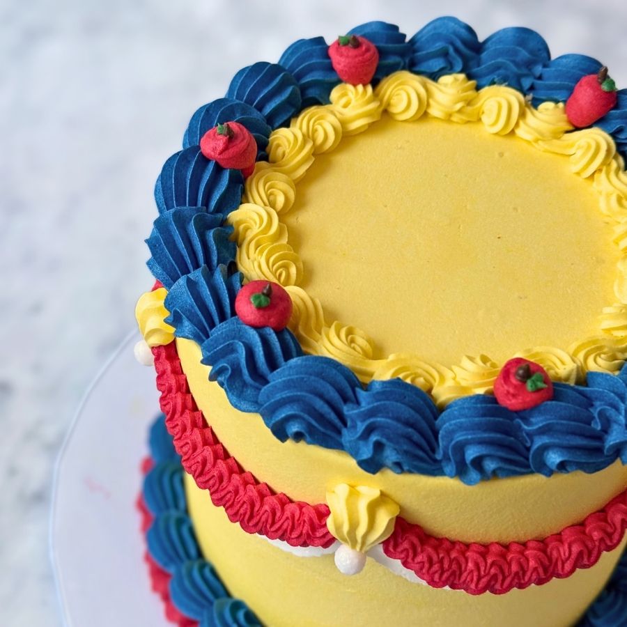 A close-up side view of a cake with deep blue frosting. It is decorated with red frosting swags, gold beads, and a large red apple decoration. White frosting ruffles line the top and bottom.