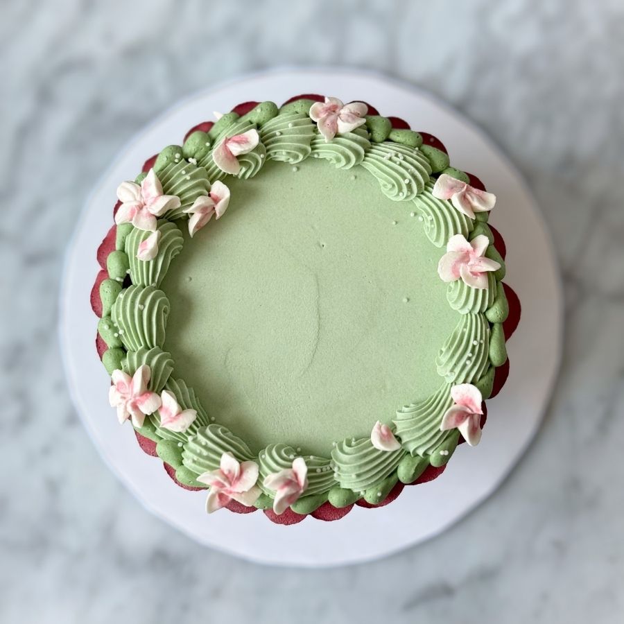 An overhead view of a round cake frosted in smooth sage green buttercream. The top edge is decorated with a crown of textured green frosting swirls and several piped pink blossoms. A deep red scalloped border is visible around the outer base of the cake.