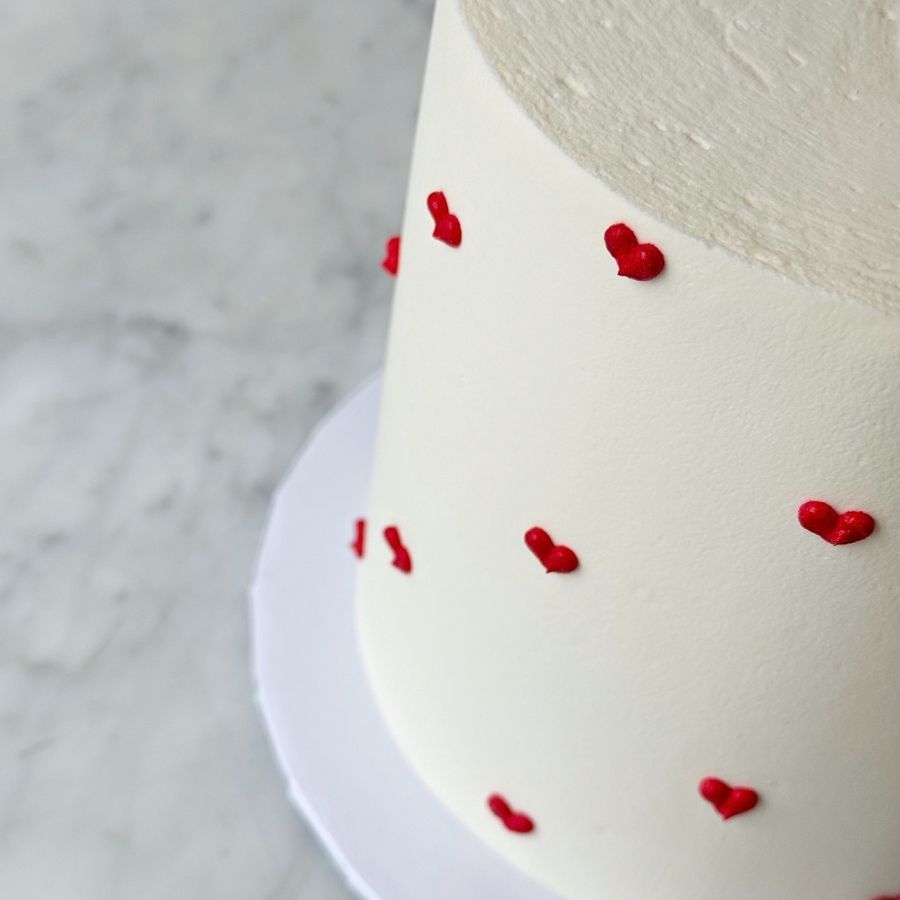 A close-up, angled shot of the top edge of a minimalist cake. The image showcases the smooth texture of the white frosting and the raised detail of small, vibrant red buttercream hearts piped onto the side. The lighting is soft and bright, emphasizing the clean design.