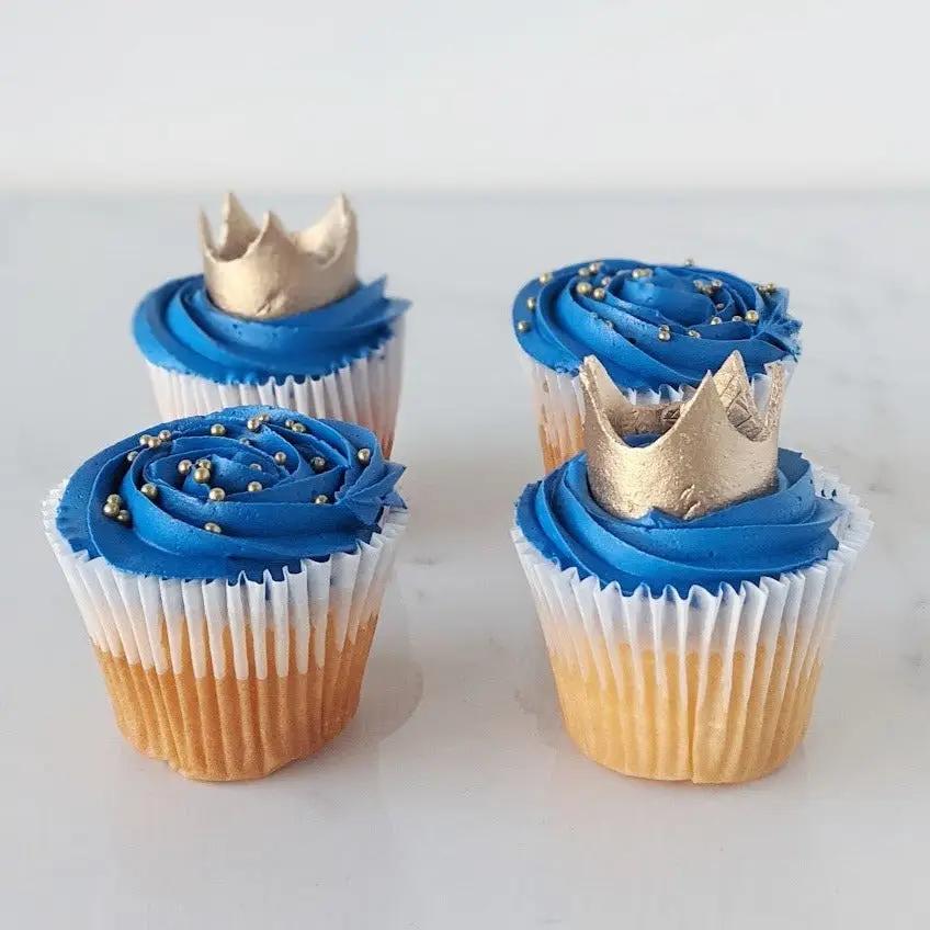Four cupcakes with blue frosting and, small gold balls and crown decorations.