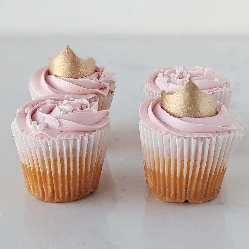 Four cupcakes with pink frosting and small gold crown decorations.