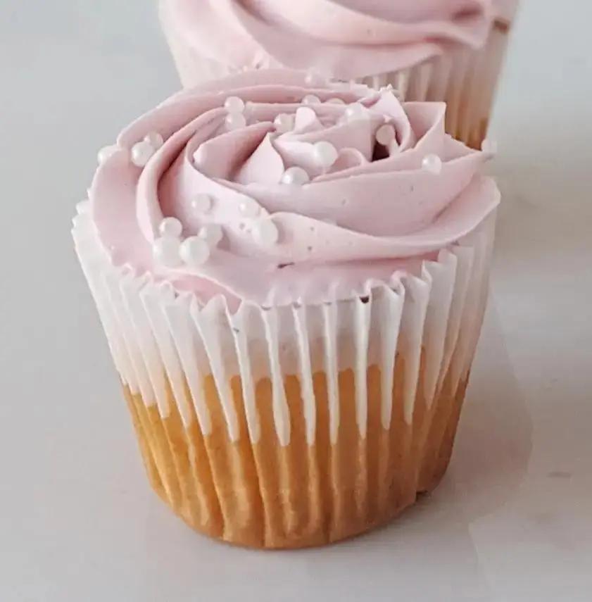 Two cupcakes, one in the foreground and one slightly behind, with light pink frosting swirled on top. The cupcakes are in light-colored paper liners.