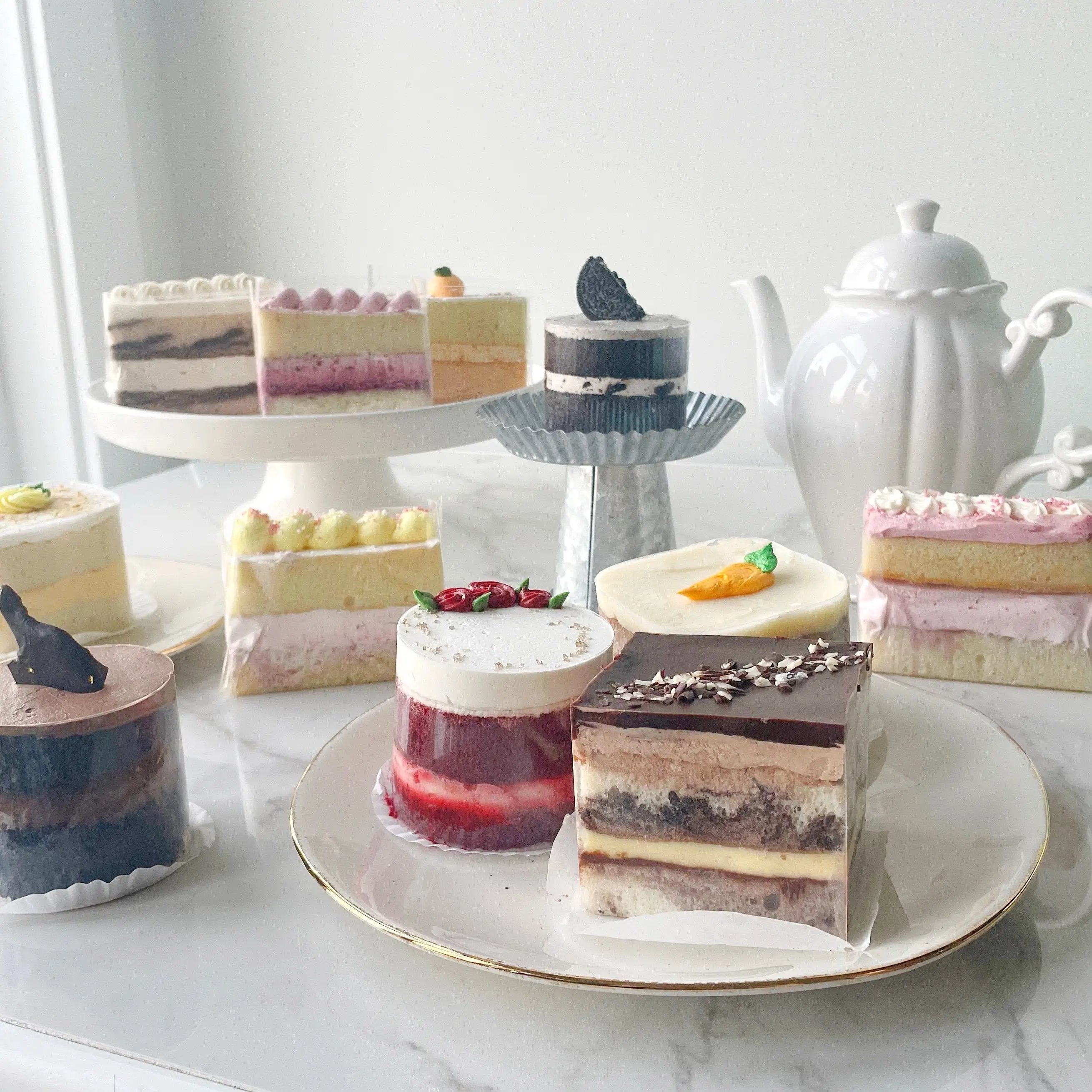 A variety of cake slices and small cakes on a table, including a strawberry cake, chocolate cake, and several other colorful flavors.
