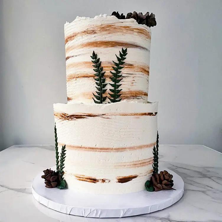 A two-tiered round wedding cake with a wood-like appearance and greenery decorations and brown frosting as pinecone.