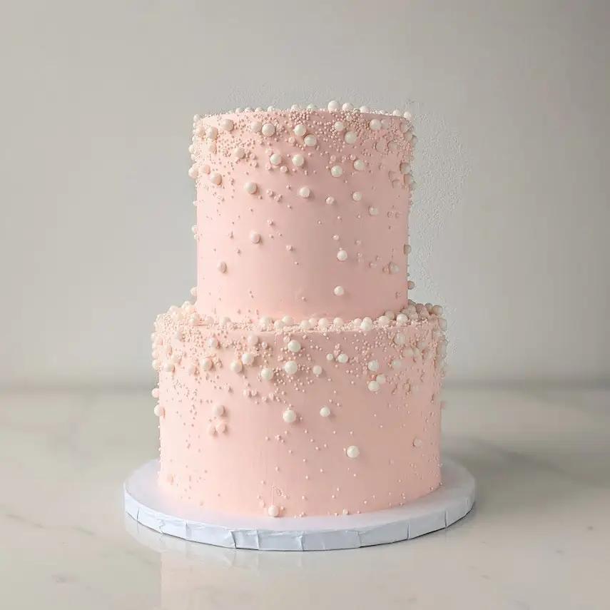 A two-tiered cake with light pink frosting. The tiers are decorated with scattered, small, white edible pearls.