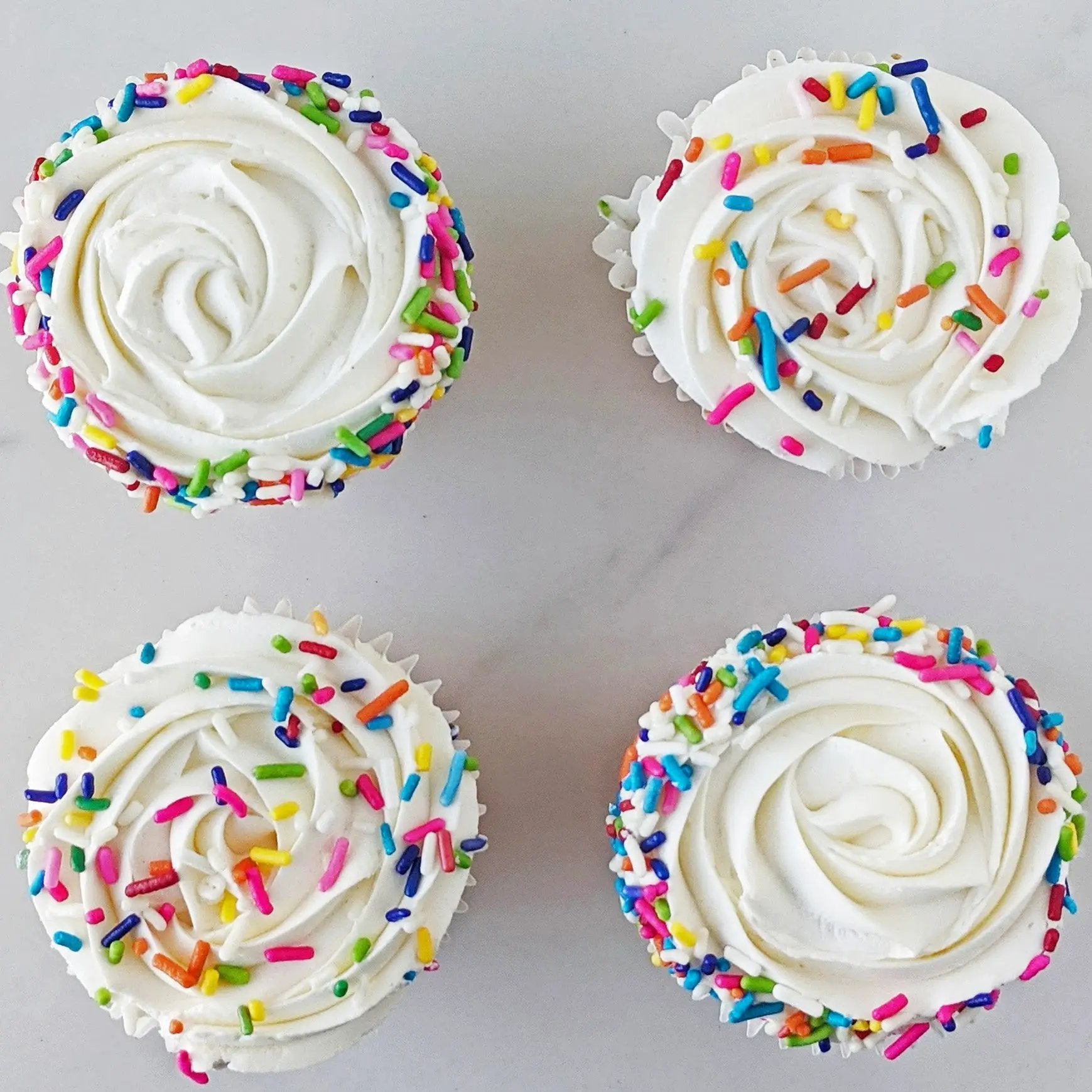 A set of four cupcakes with white frosting and multicolored sprinkles on top.
