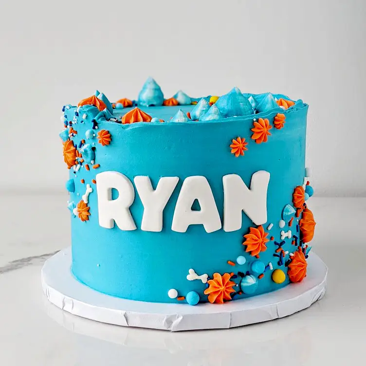 A birthday cake with blue icing and orange and white decorative elements, topped with the name 'RYAN' in white letters.